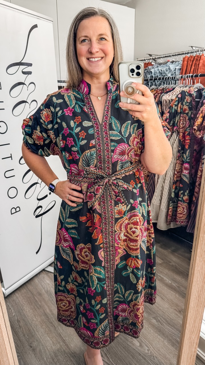 Woman with shoulder-length hair taking a mirror selfie in a clothing store, wearing a colorful patterned dress.