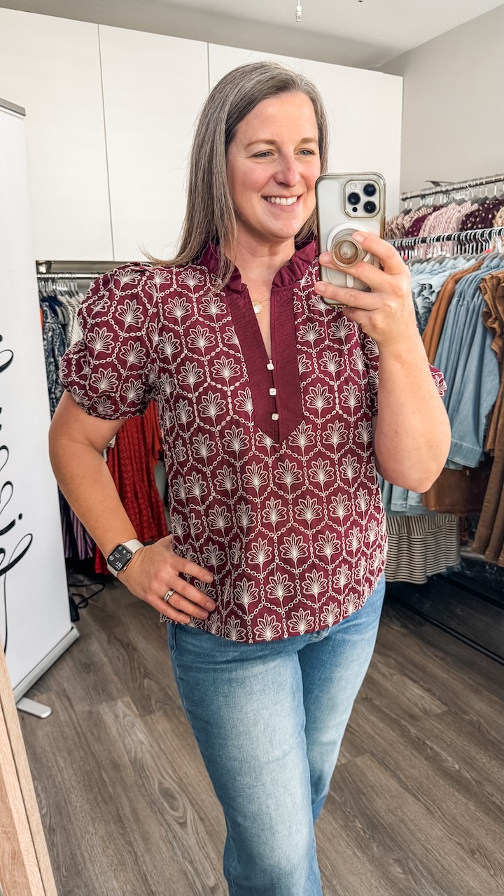 Woman taking a mirror selfie, smiling, wearing a patterned red blouse and jeans, in a clothing store fitting room.