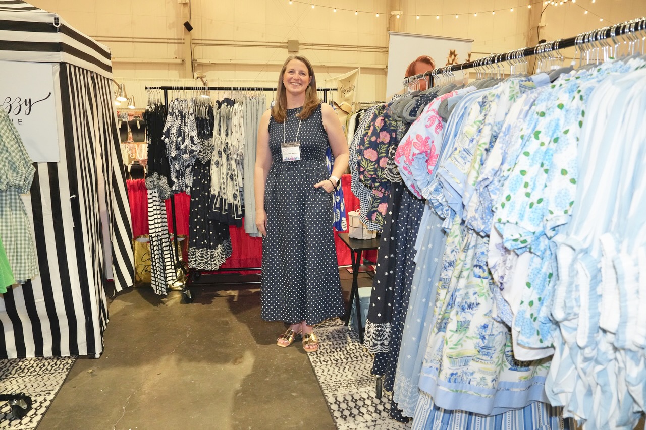 Woman standing in clothing store aisle with racks of dresses on both sides, smiling, wearing a sleeveless dress.