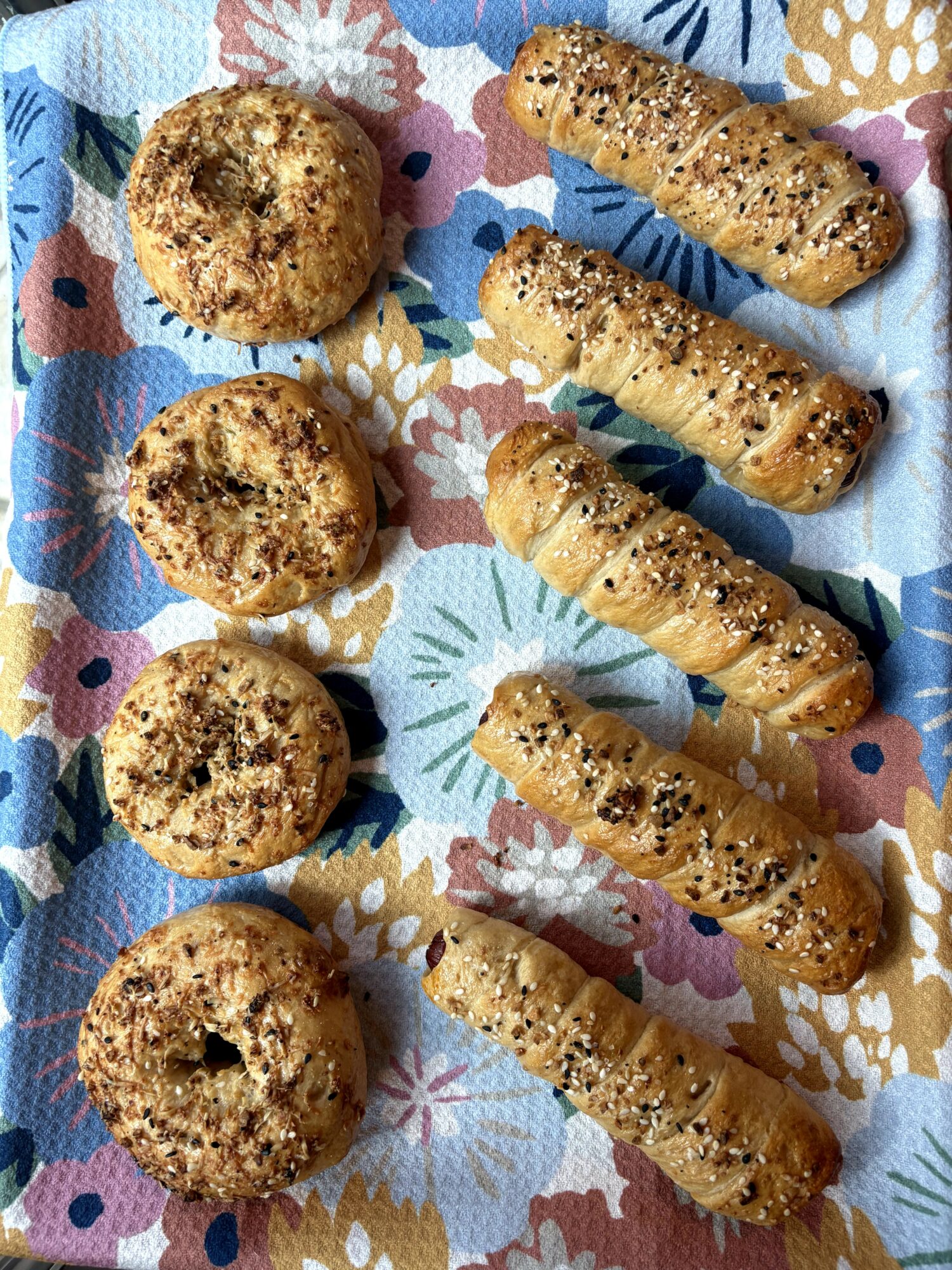 Six breaded, fried balls and five breadsticks with sesame seeds on a floral-patterned cloth.