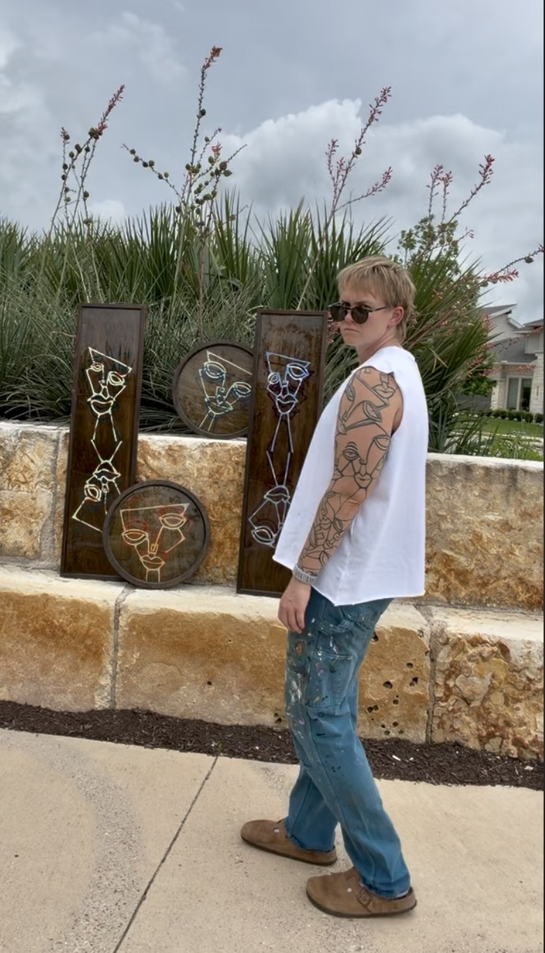 Person with tattoos and sunglasses standing outdoors near decorative metal art pieces, plants, and stone wall.