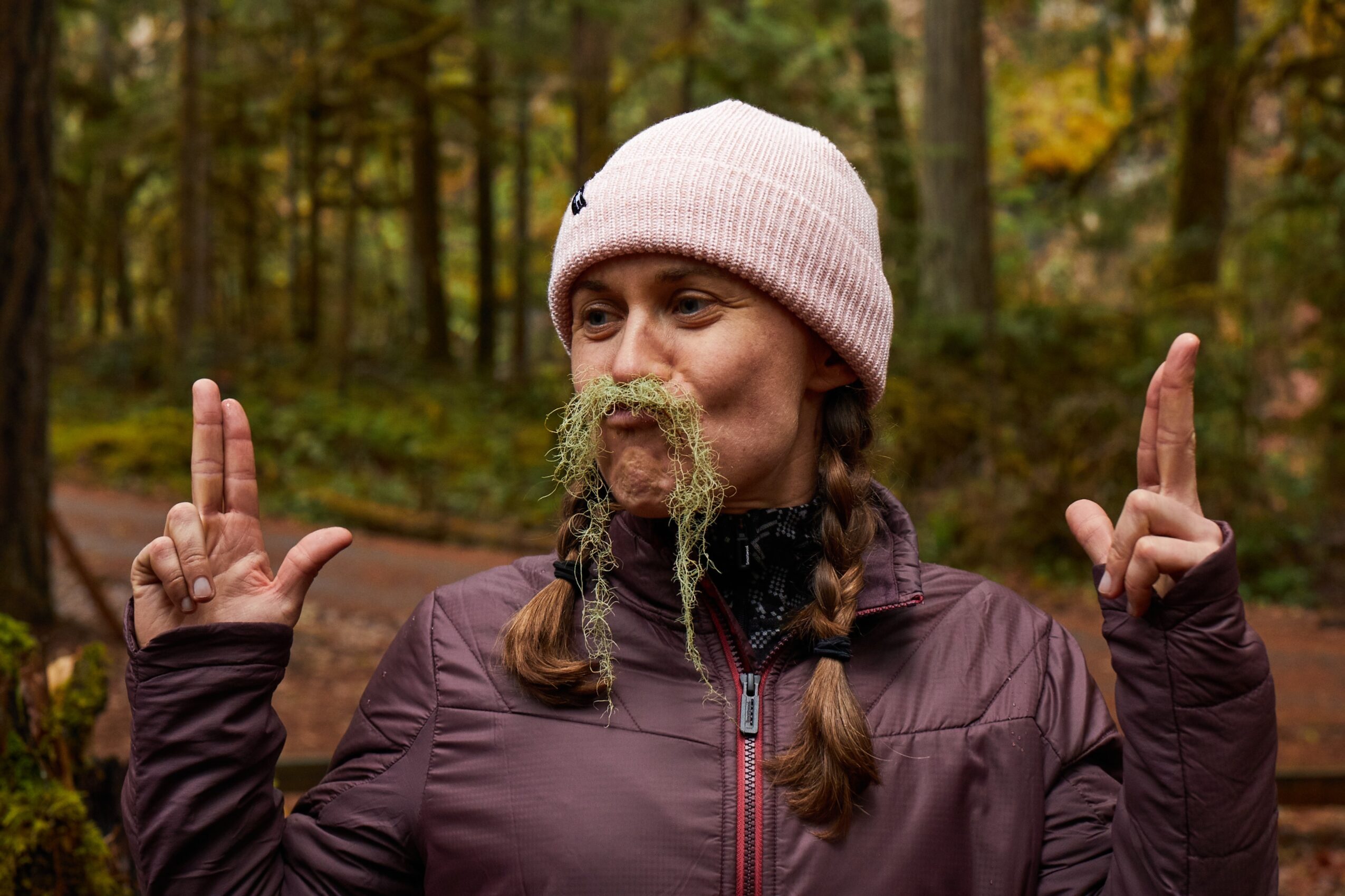 Person in a pink knit hat stands in a wooded area with plants hanging from their mouth, holding up both hands.
