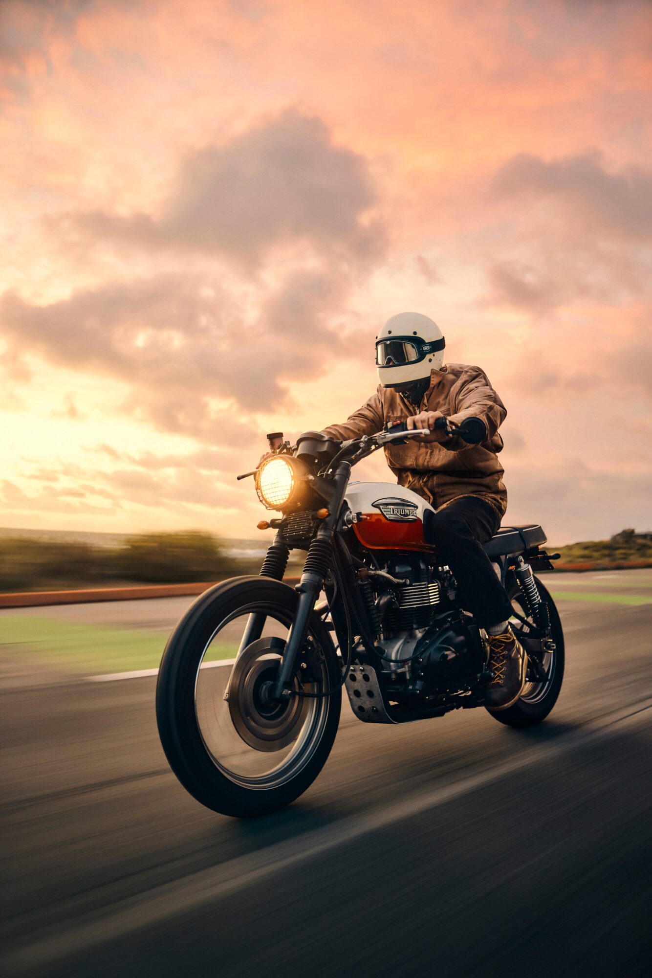 Helmeted rider on a motorcycle traveling along a road beneath a cloudy sunset sky.