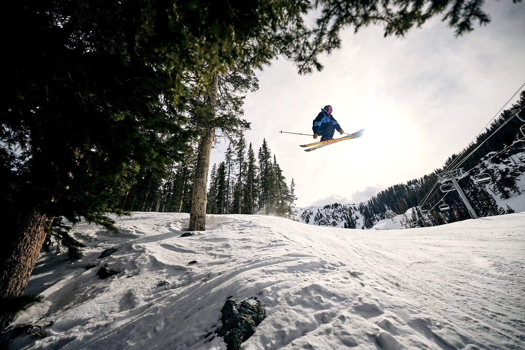 A skier jumps above a snowy slope, framed by trees and ski-lift equipment.