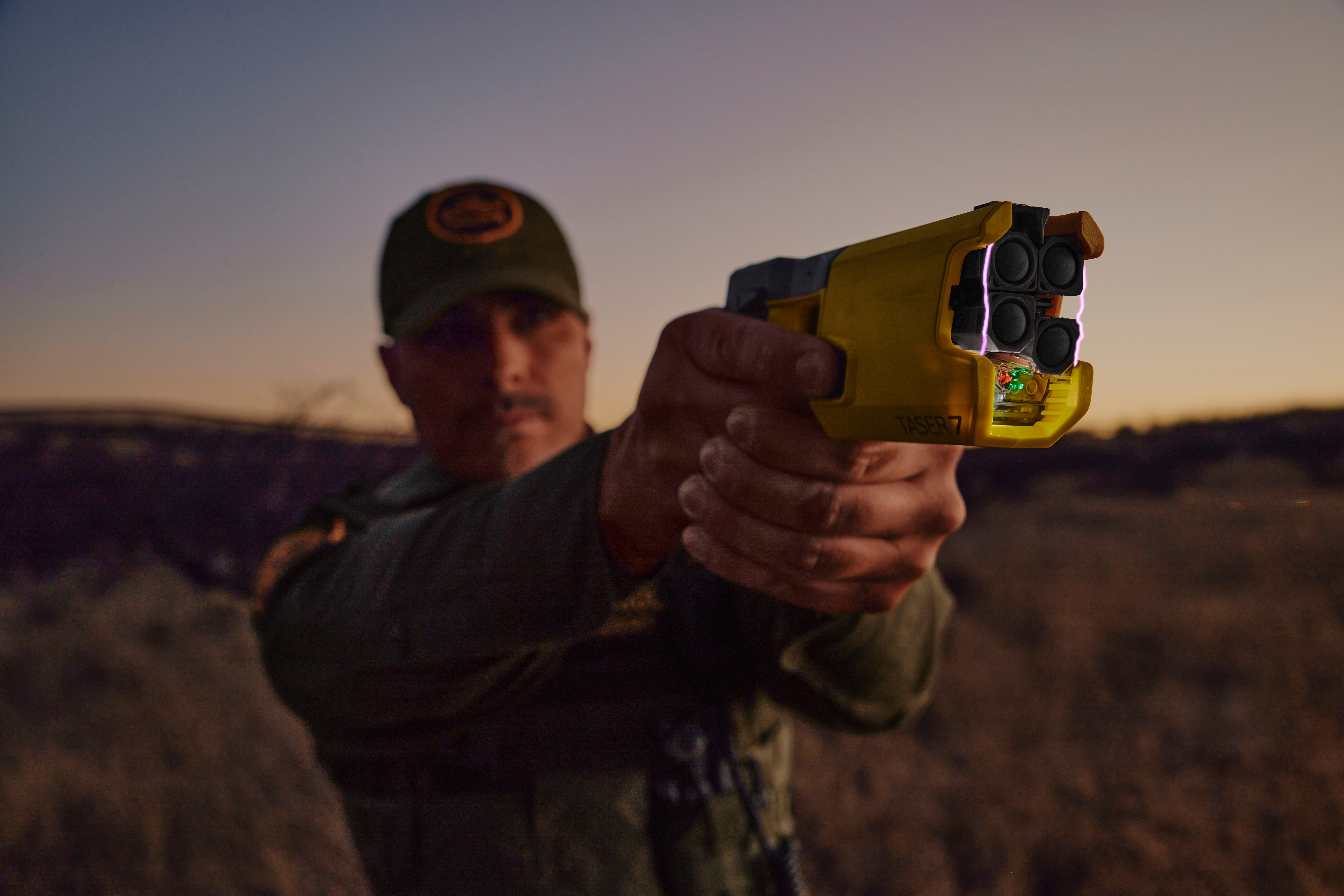 Person wearing a cap aims a yellow handheld device toward the camera in an open landscape.