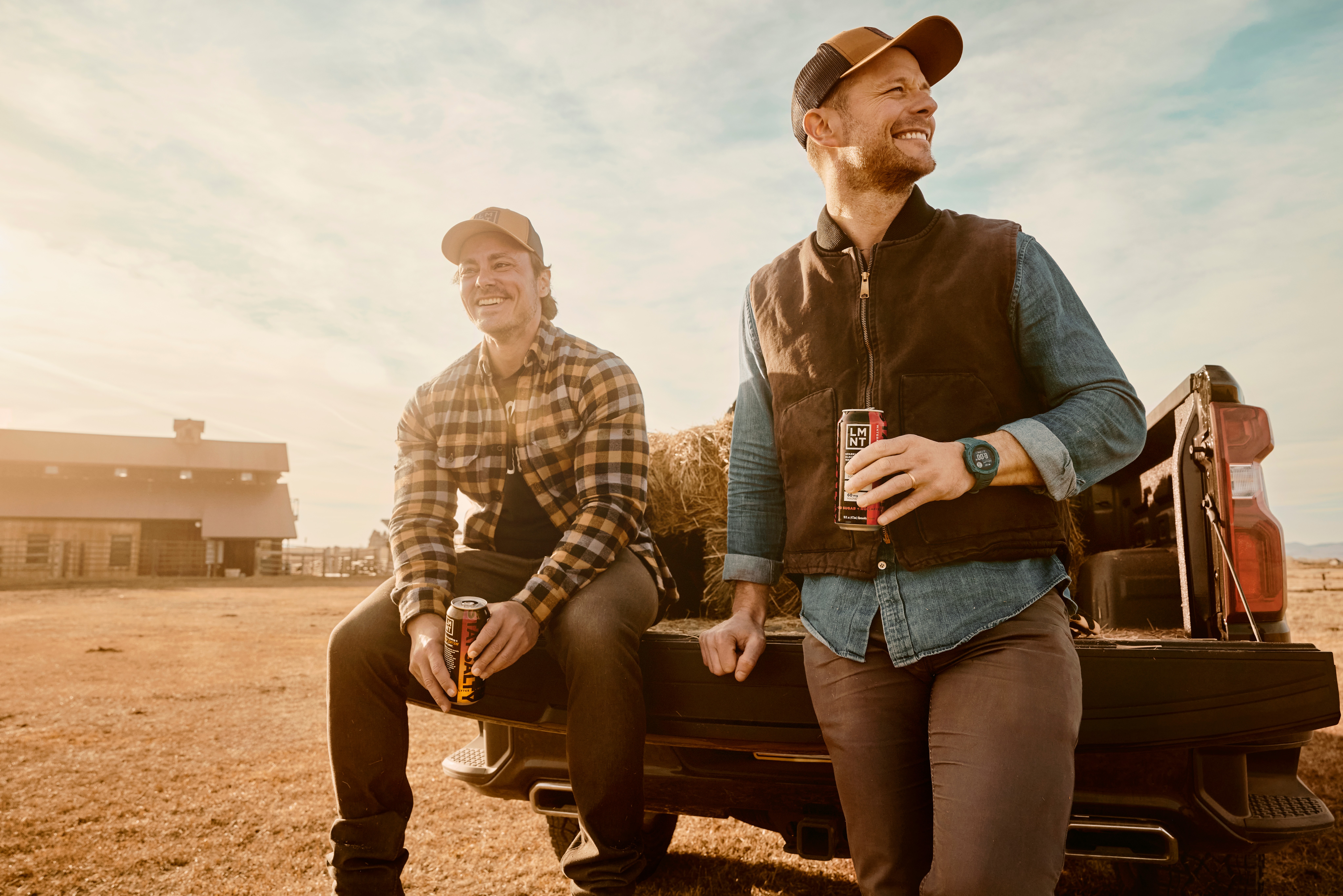 Two people sit on the back of a pickup truck in a dry field, with a building and equipment nearby.