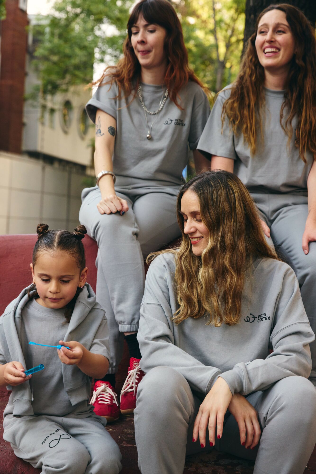 Four people sitting outdoors, smiling, wearing matching gray sweatshirts, with trees and a building in the background.