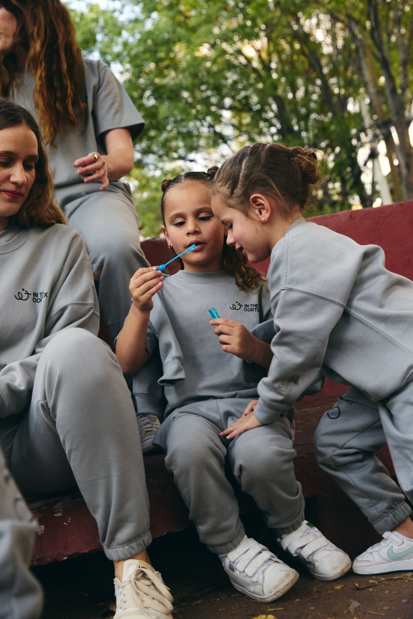 Group of children sitting outdoors, some children are talking and holding objects, with trees in the background.