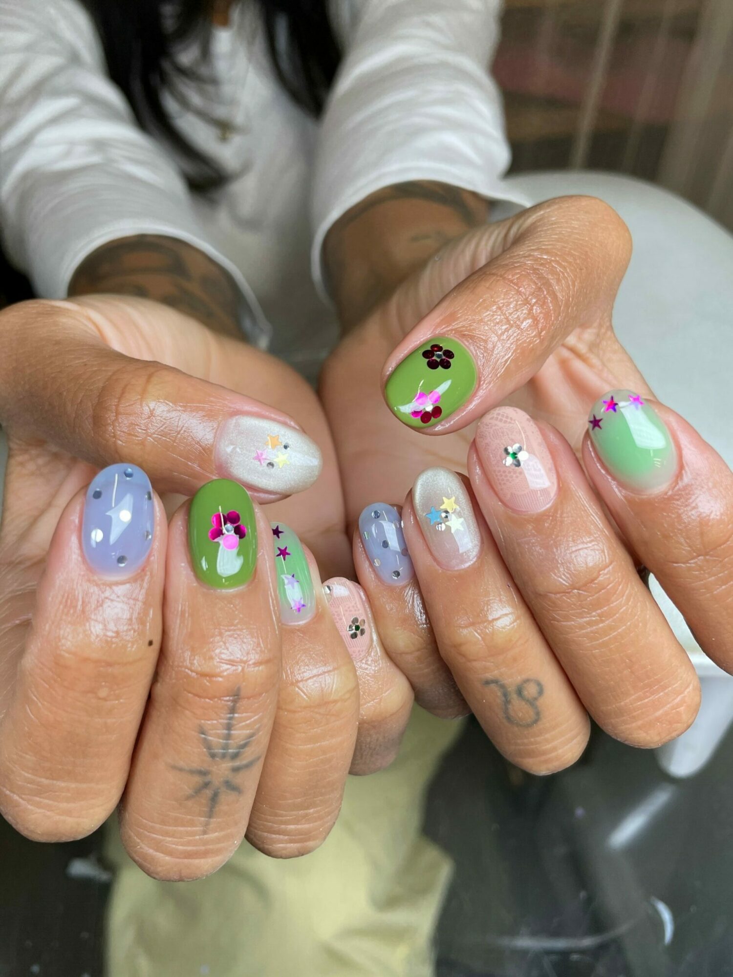 Close-up of hands with colorful, decorated nails featuring flowers, clouds, and pastel colors, on a reflective surface.