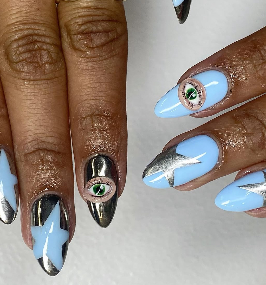 Close-up of hands with blue, black, and silver nail art, including eye and star designs, on a white background.
