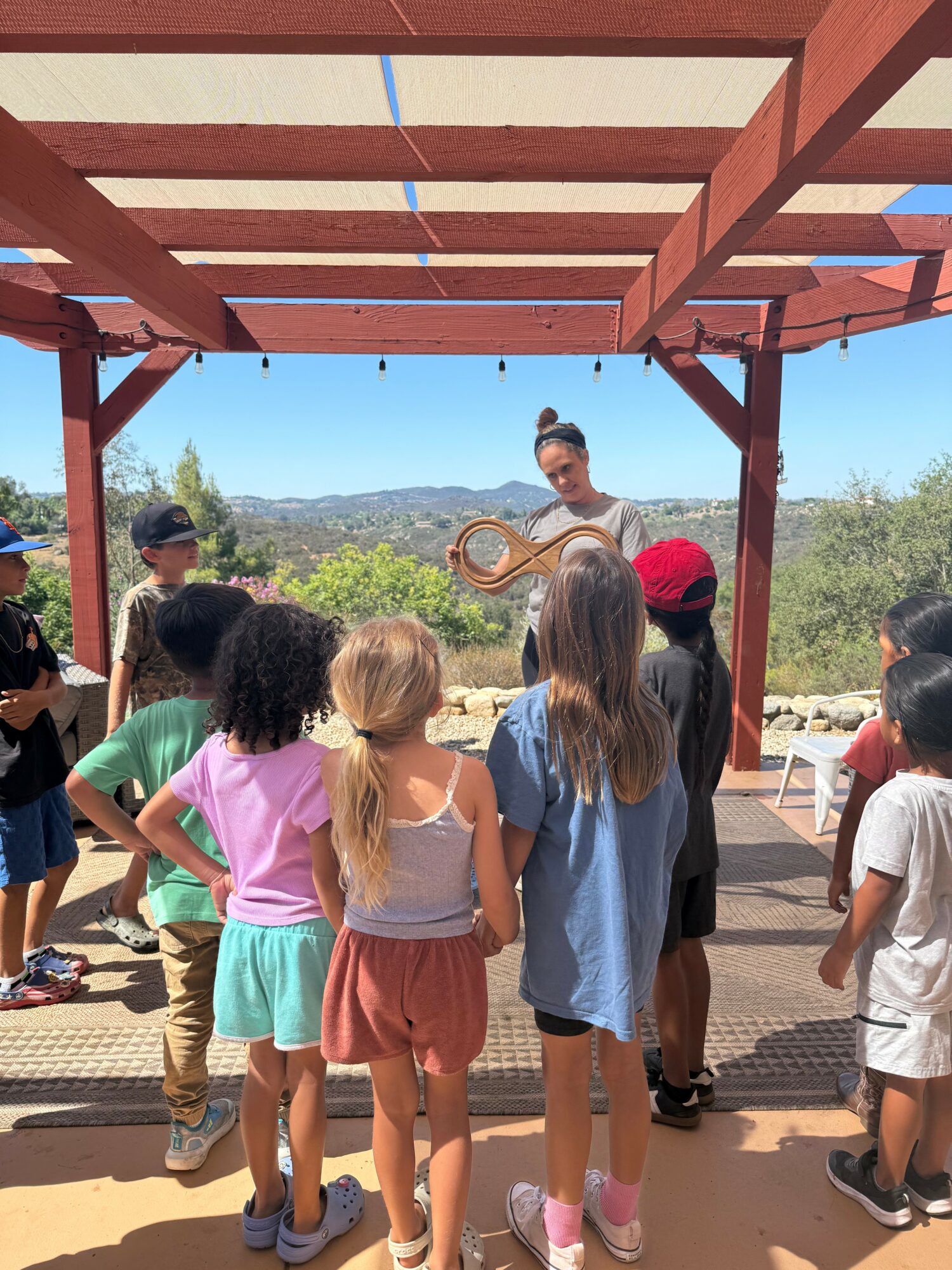 Group of children gathered around a woman holding a wooden object outdoors under a wooden pergola, with trees and mountains in the background.