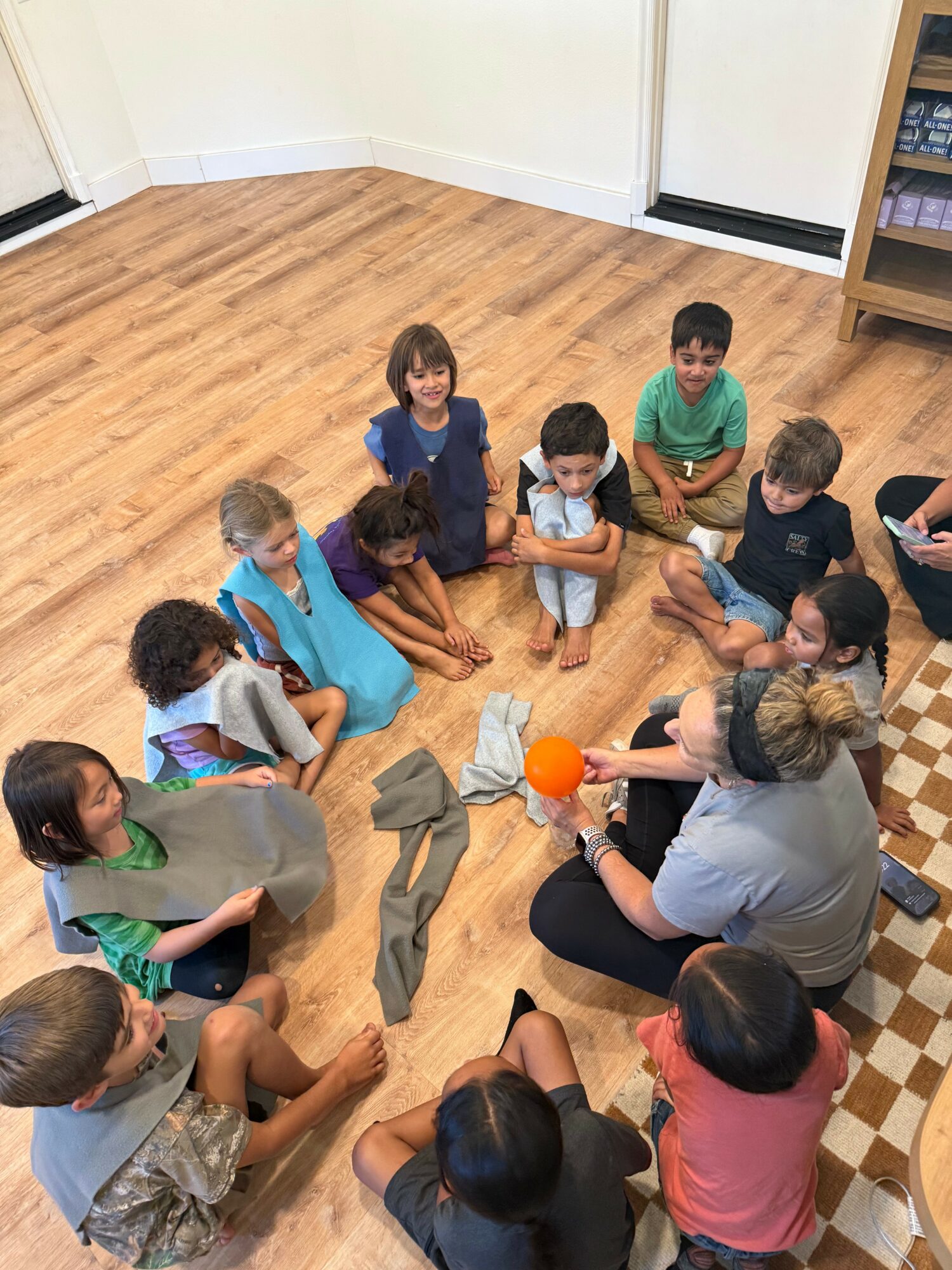 Children sitting in a circle on a wooden floor, engaging in a group activity with a woman holding an orange ball.