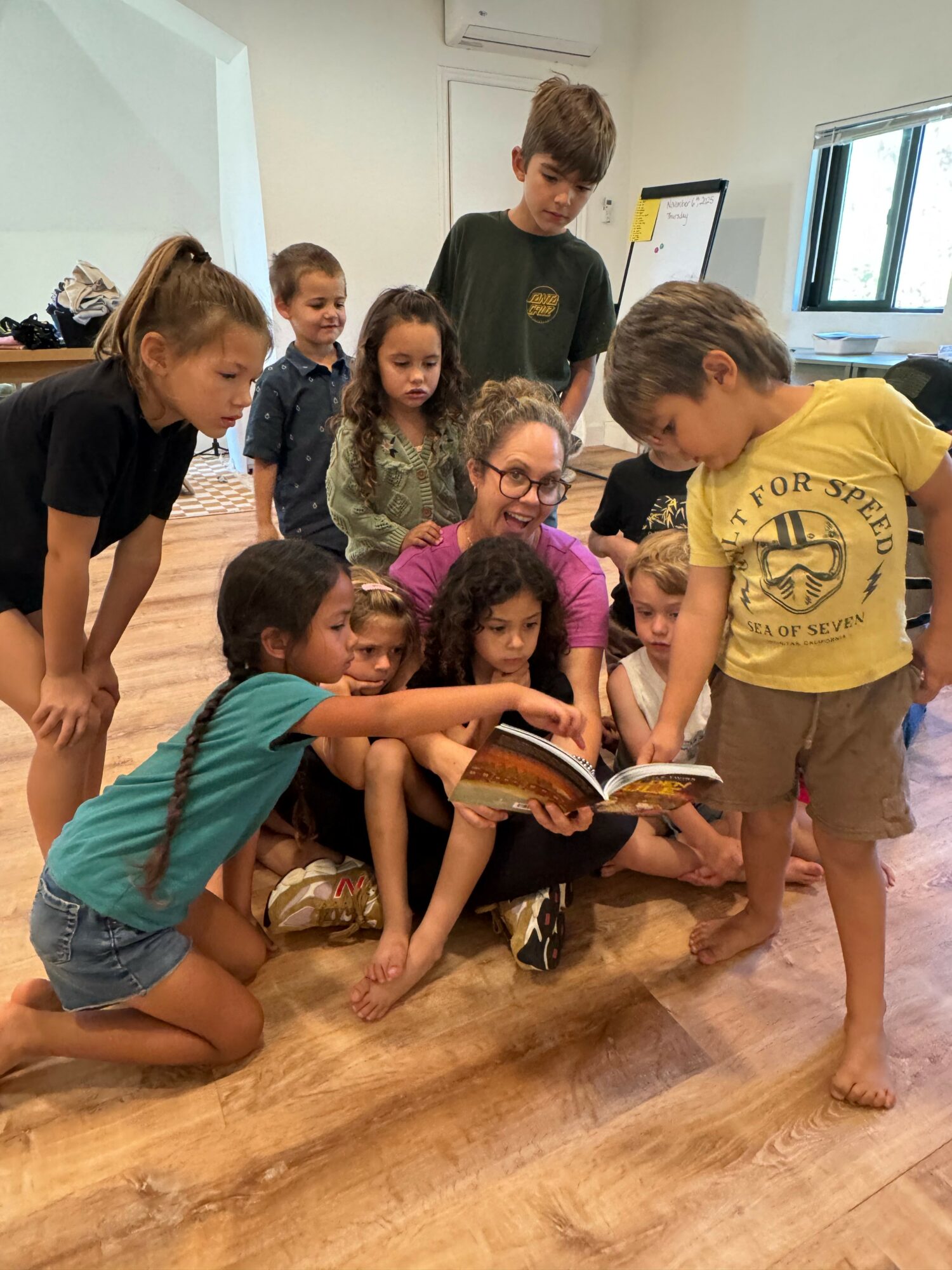 Group of children gathered around a woman reading a book on the floor in a classroom, with some children sitting and others standing.