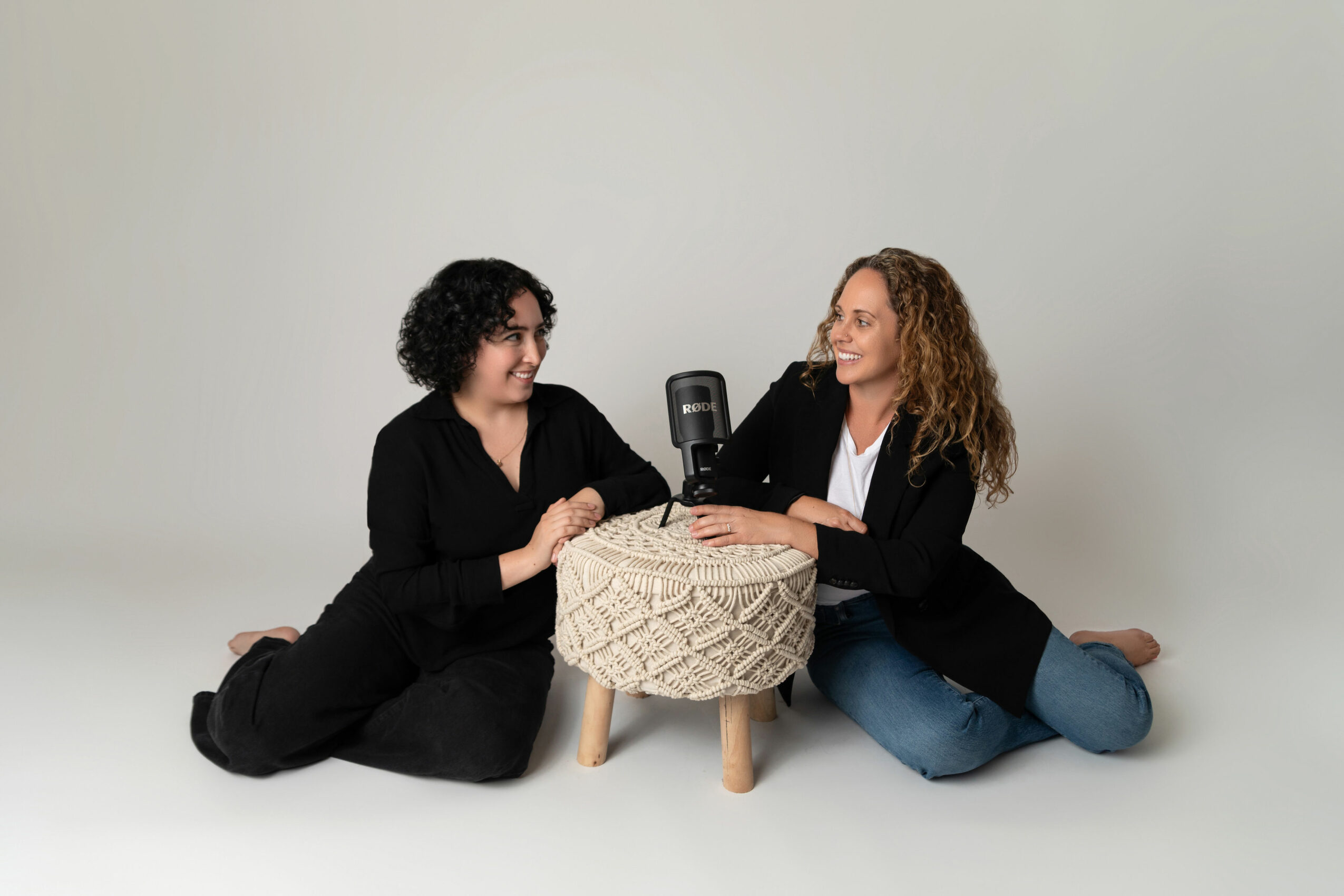 Two women sit on the floor facing each other, smiling, with a small round table between them, against a plain background.