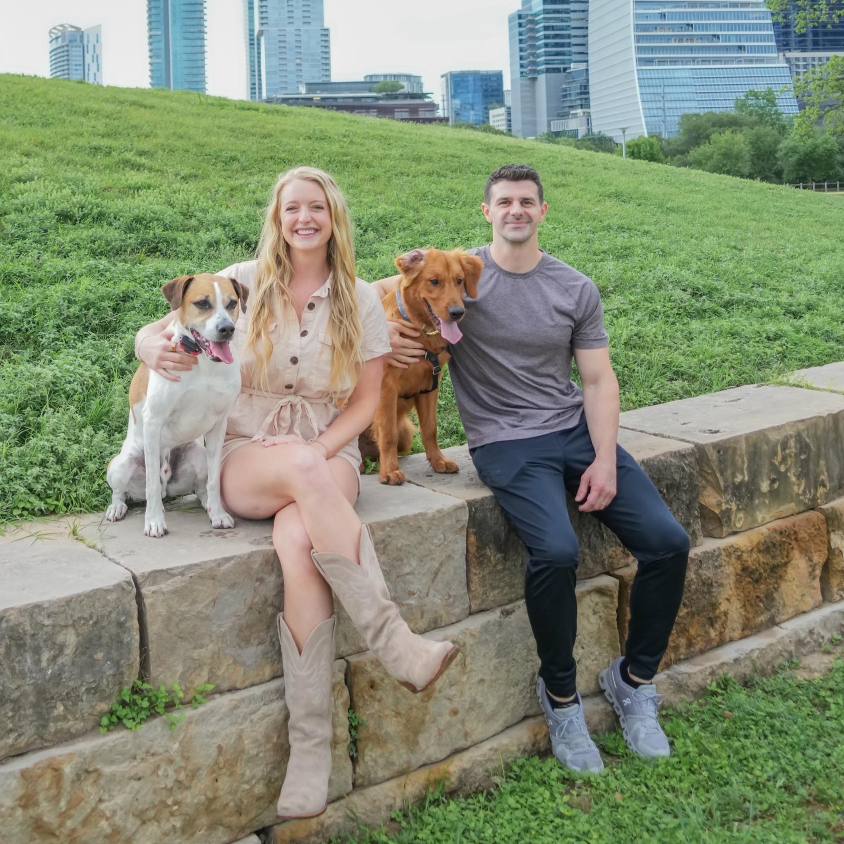 A smiling woman and man sit on a stone wall with two dogs in a park, city skyline in background.