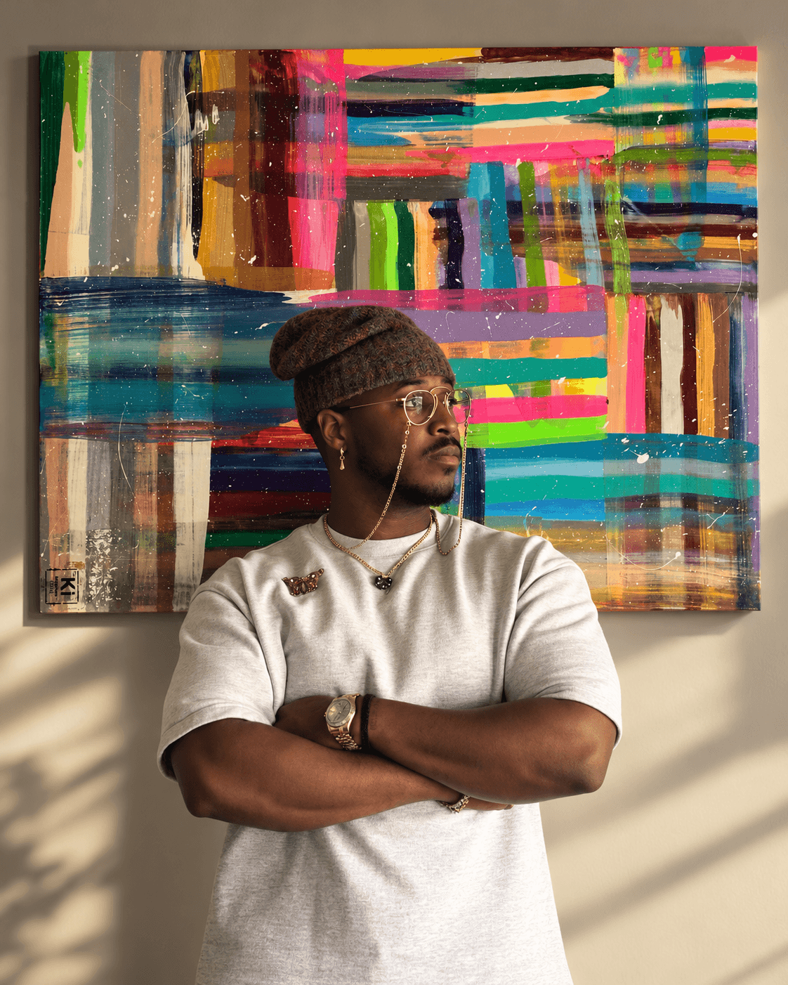Man with glasses, wearing a beanie and white shirt, stands with arms crossed in front of colorful abstract painting.