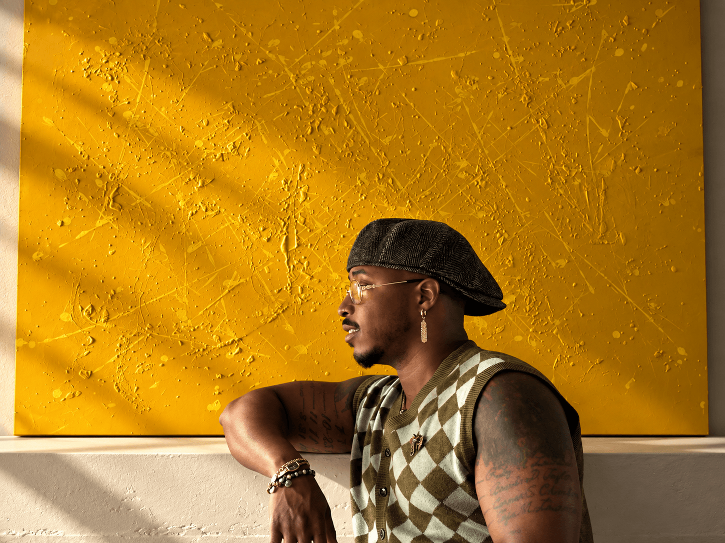Man with glasses, wearing a cap backwards and a sleeveless argyle shirt, leaning on a white surface against a yellow textured background.