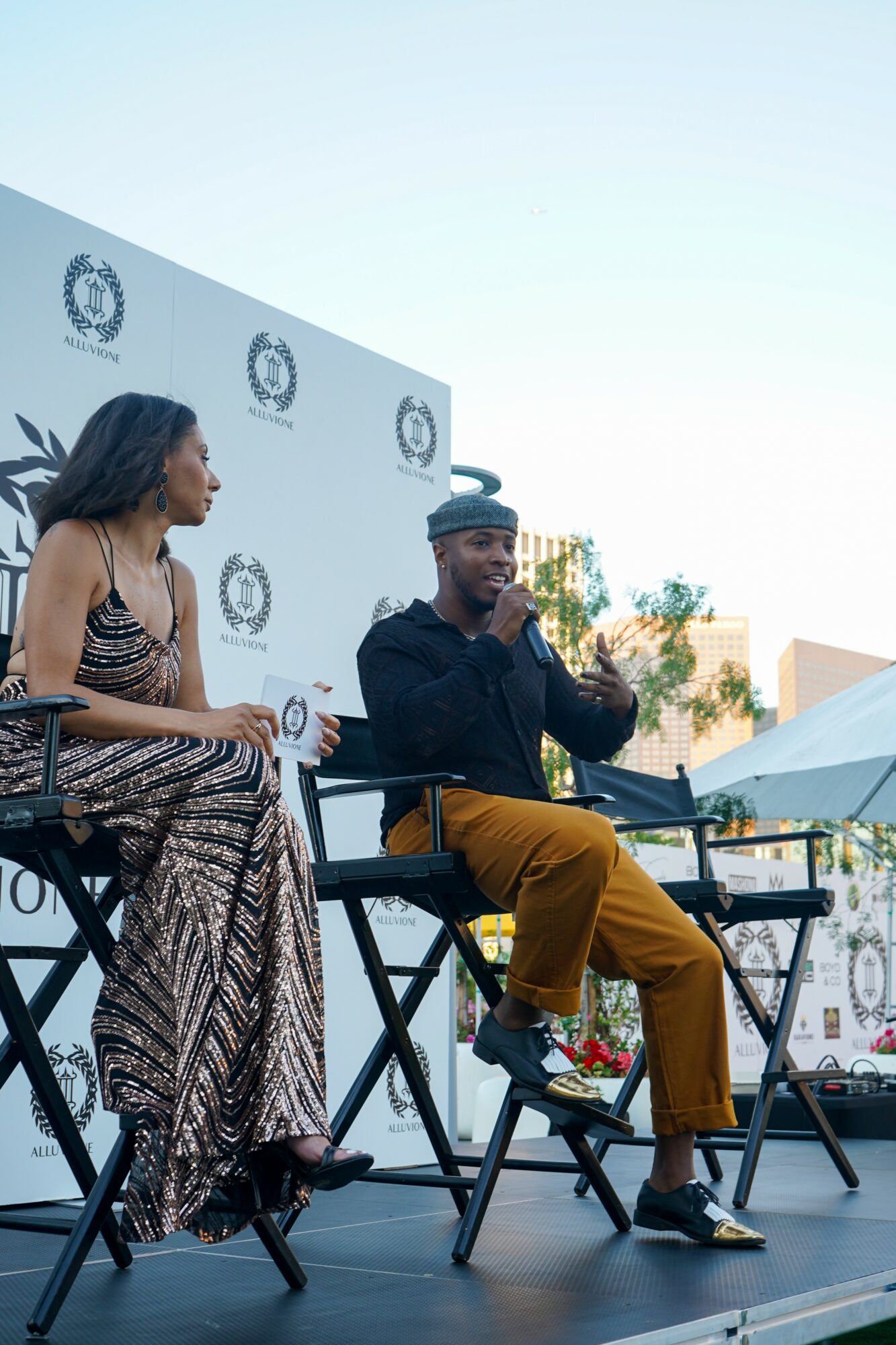 Two people sitting on stage, one clapping, with a backdrop displaying logos, outdoors with trees and tents nearby.