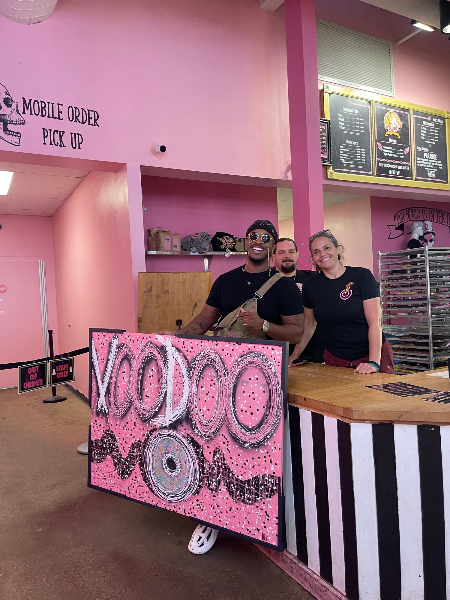 Three people behind a counter in a pink-themed shop, with a pink and black display in front, and menu boards on the wall.