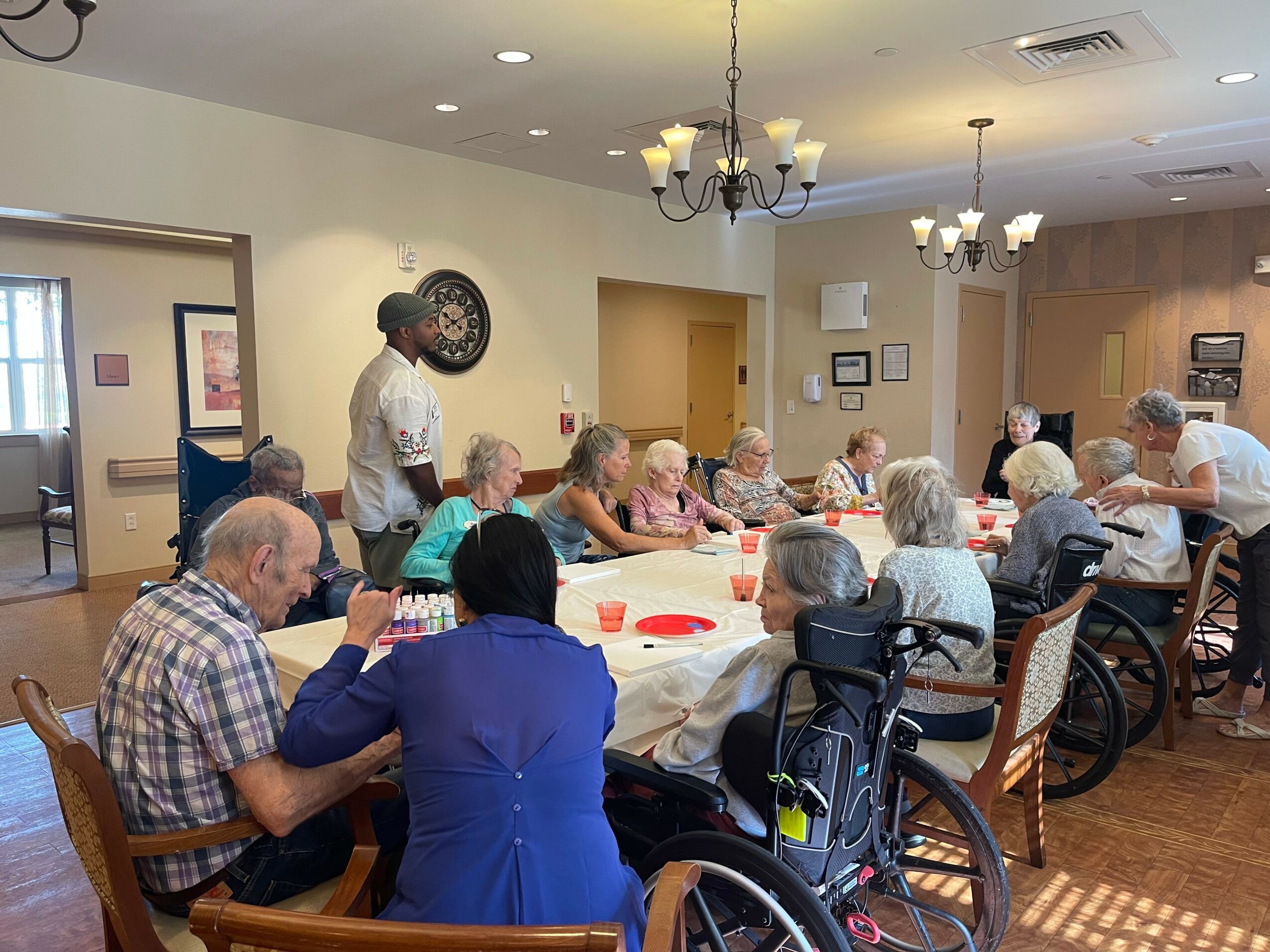Group of elderly people sitting around a table in a dining room, some in wheelchairs, with staff assisting, in a care facility.