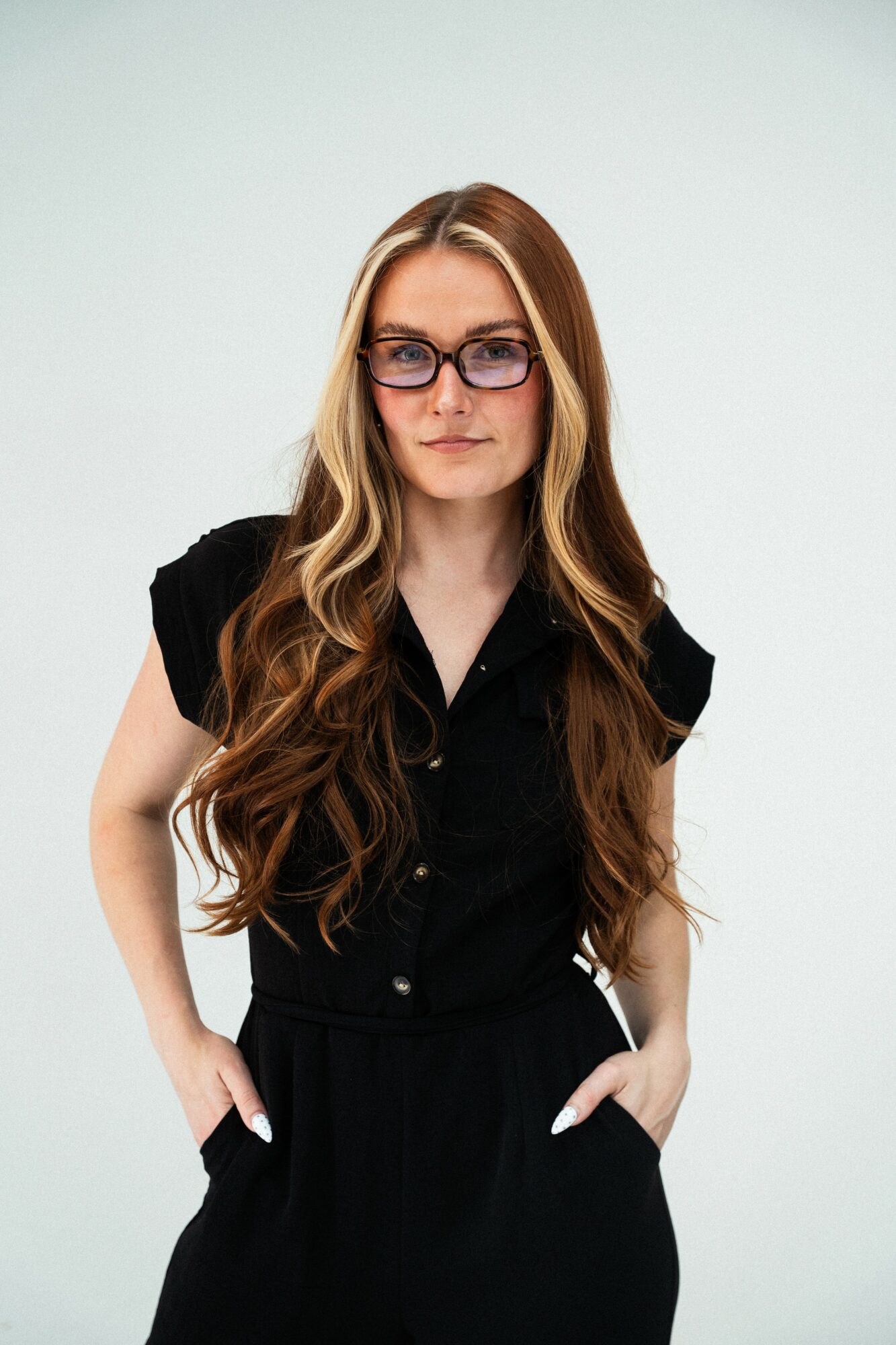 Woman with long wavy hair wearing glasses and a black outfit, standing with hands in pockets against a plain background.