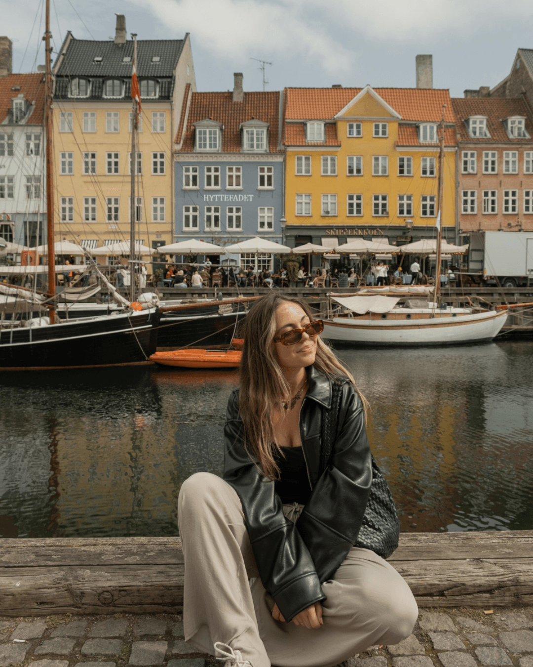 Woman sitting by a harbor with colorful buildings and boats in the background, wearing sunglasses and a black jacket.