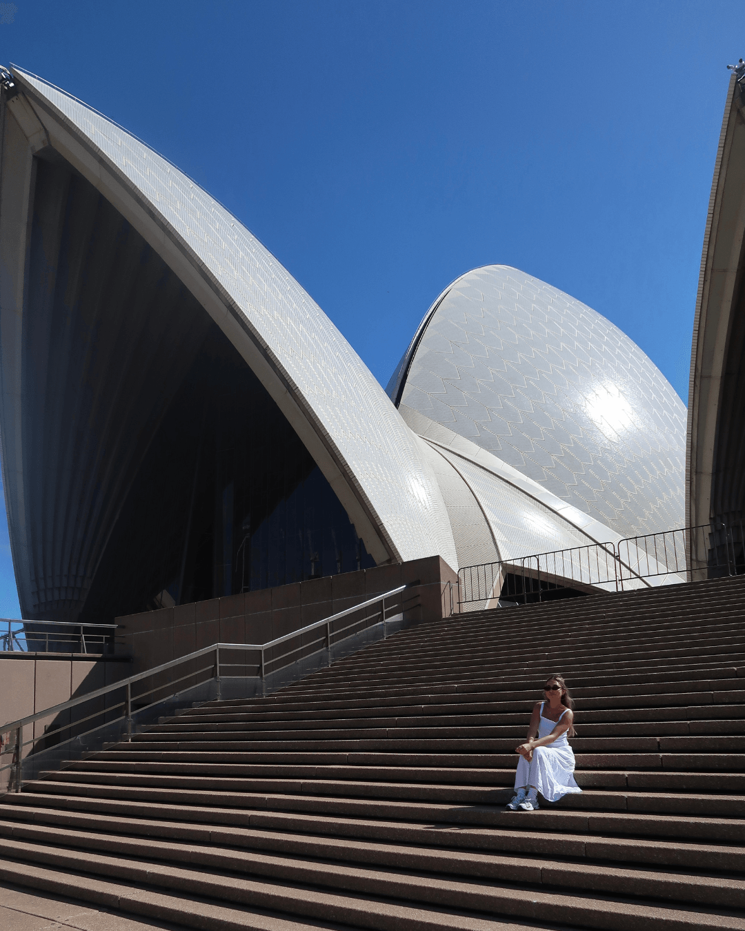 Person sitting on stairs outside Sydney Opera House with distinctive sail-like white shells against blue sky.