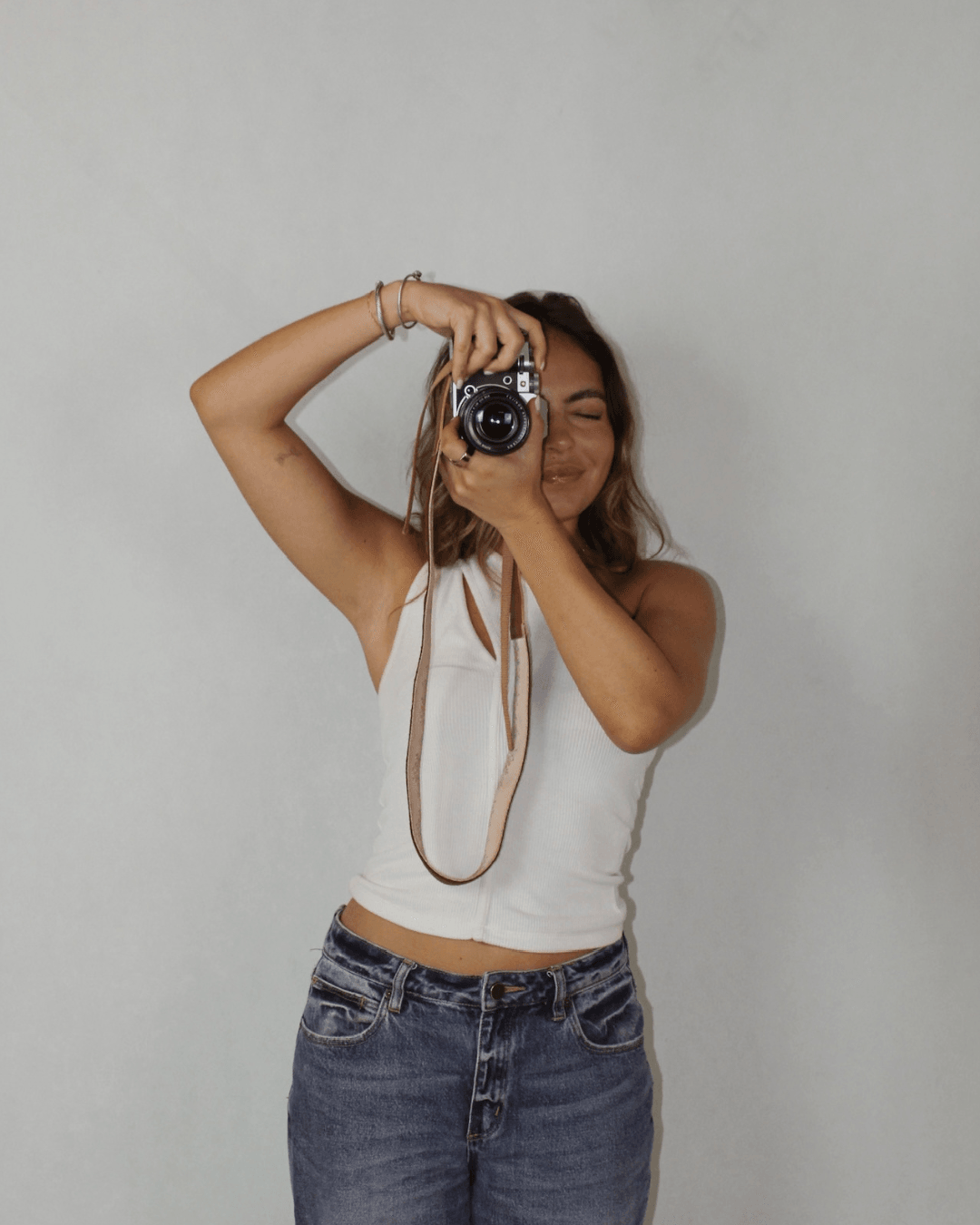Young woman taking a selfie with a camera, smiling, wearing a white tank top and jeans, standing against a plain background.