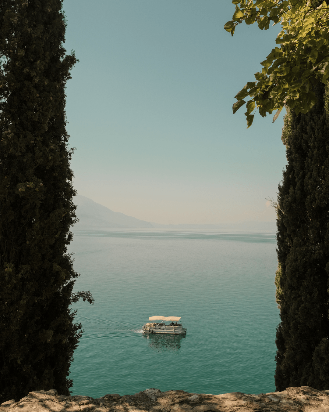 View of a boat on a calm lake framed by tall trees on both sides, with mountains in the distance and clear sky.