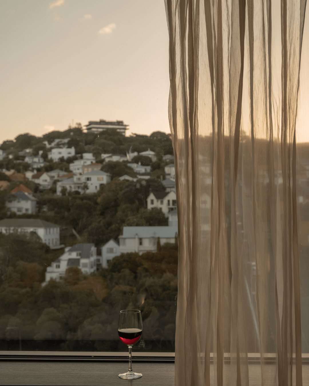 View of a hillside with white houses and trees through a window with sheer curtains, sunset lighting.