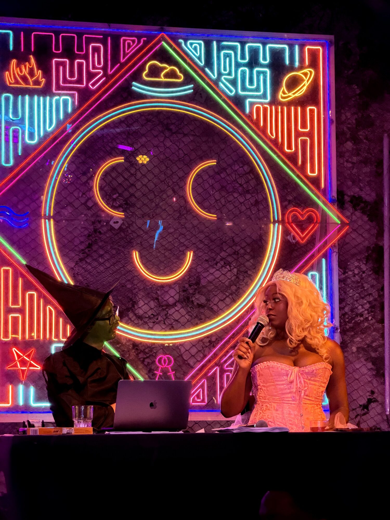 Two people on stage with a neon sign of a smiling face and various symbols behind them.