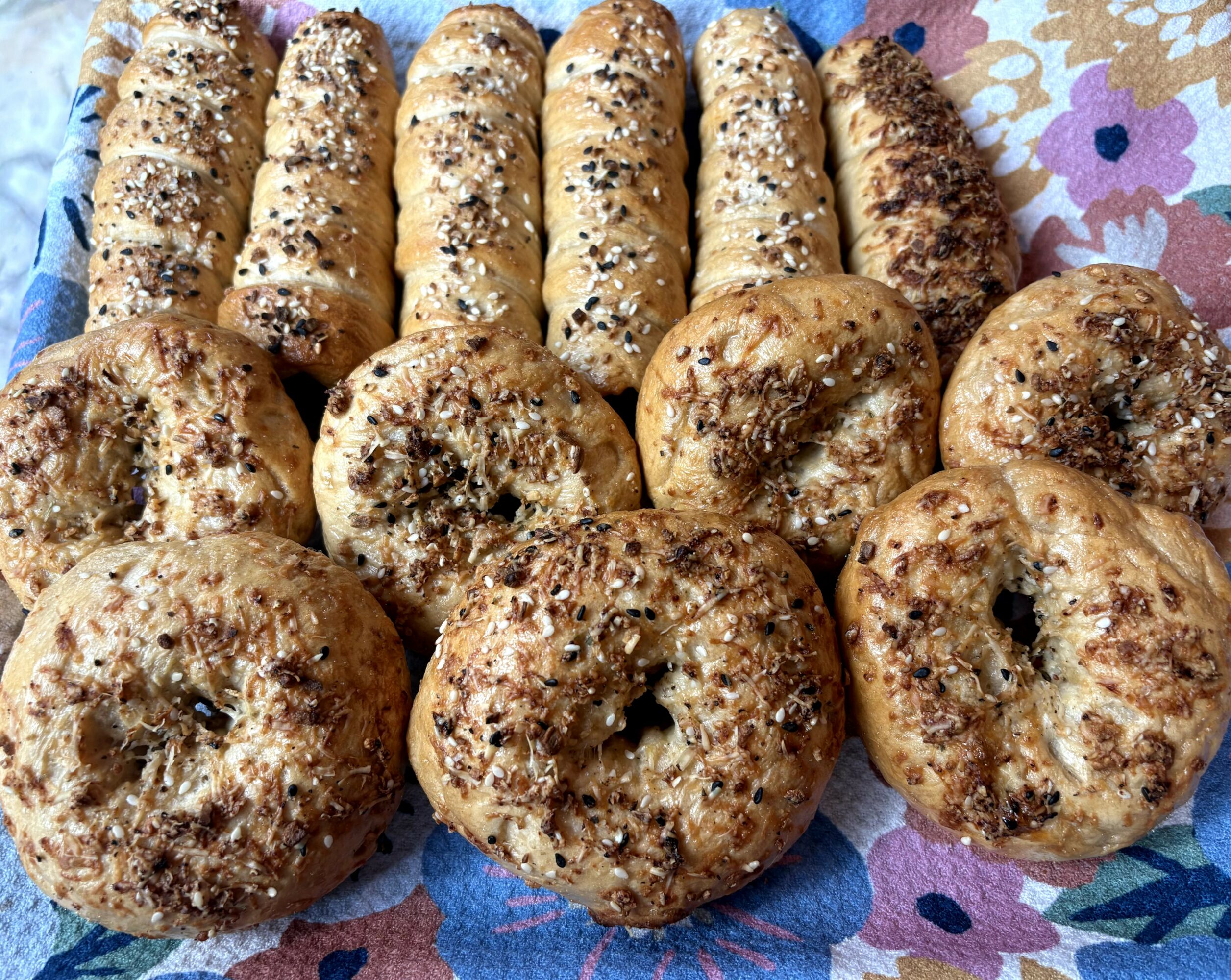 Assorted bagels with toppings on a floral cloth, some with seeds and others with chocolate or cinnamon.