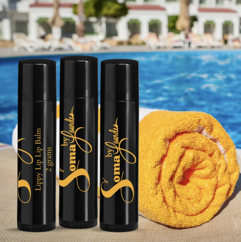 Three black lip balm sticks with gold text next to a rolled yellow towel by a poolside, with pool and lounge chairs in background.