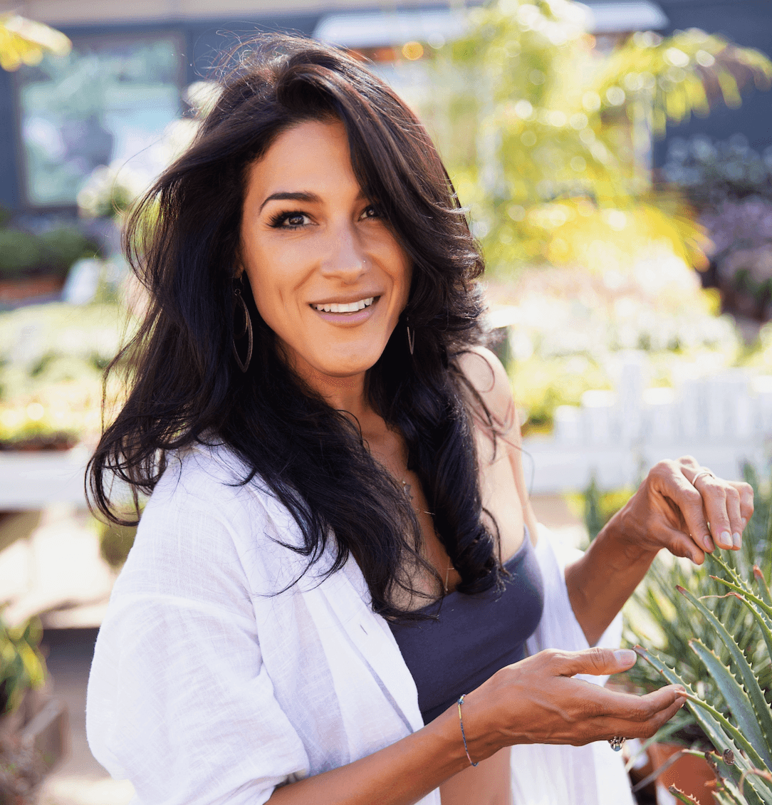 Smiling woman with dark hair in a garden, holding a plant, wearing a white shirt and dark top.