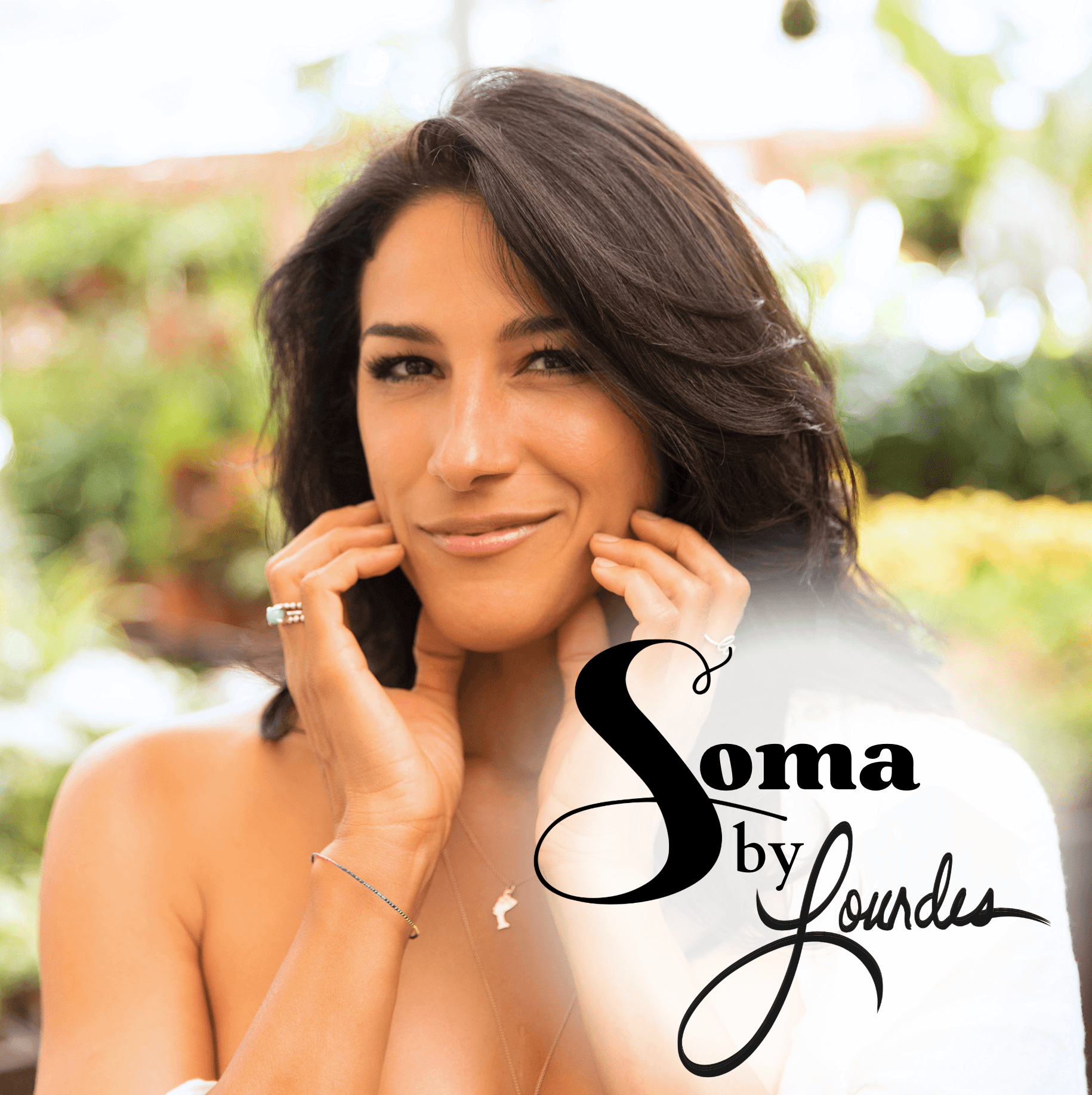 Woman with dark hair smiling outdoors, touching her face, with greenery in background.