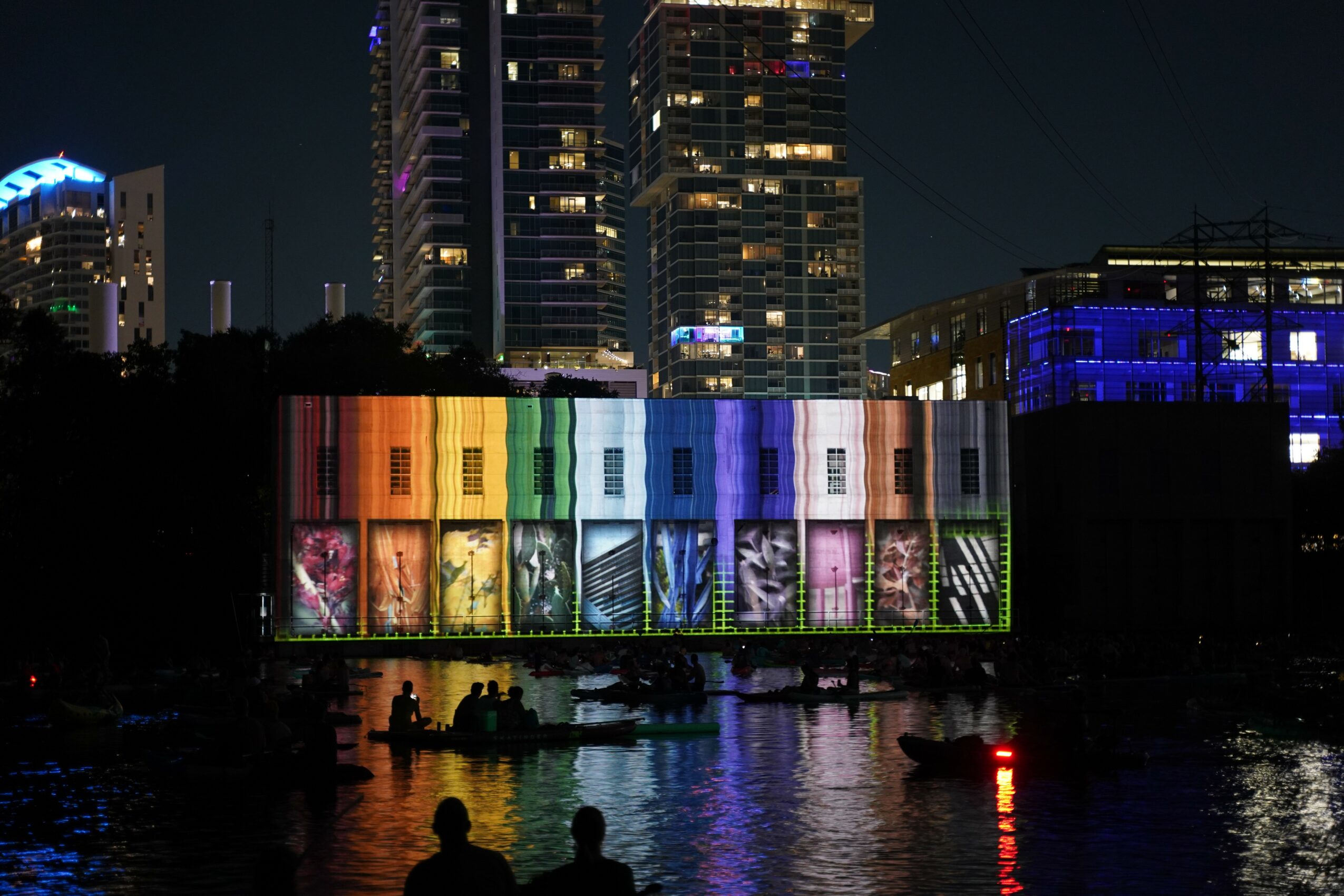 Nighttime cityscape with illuminated buildings and a colorful light display on a large screen over water, with boats and silhouettes.
