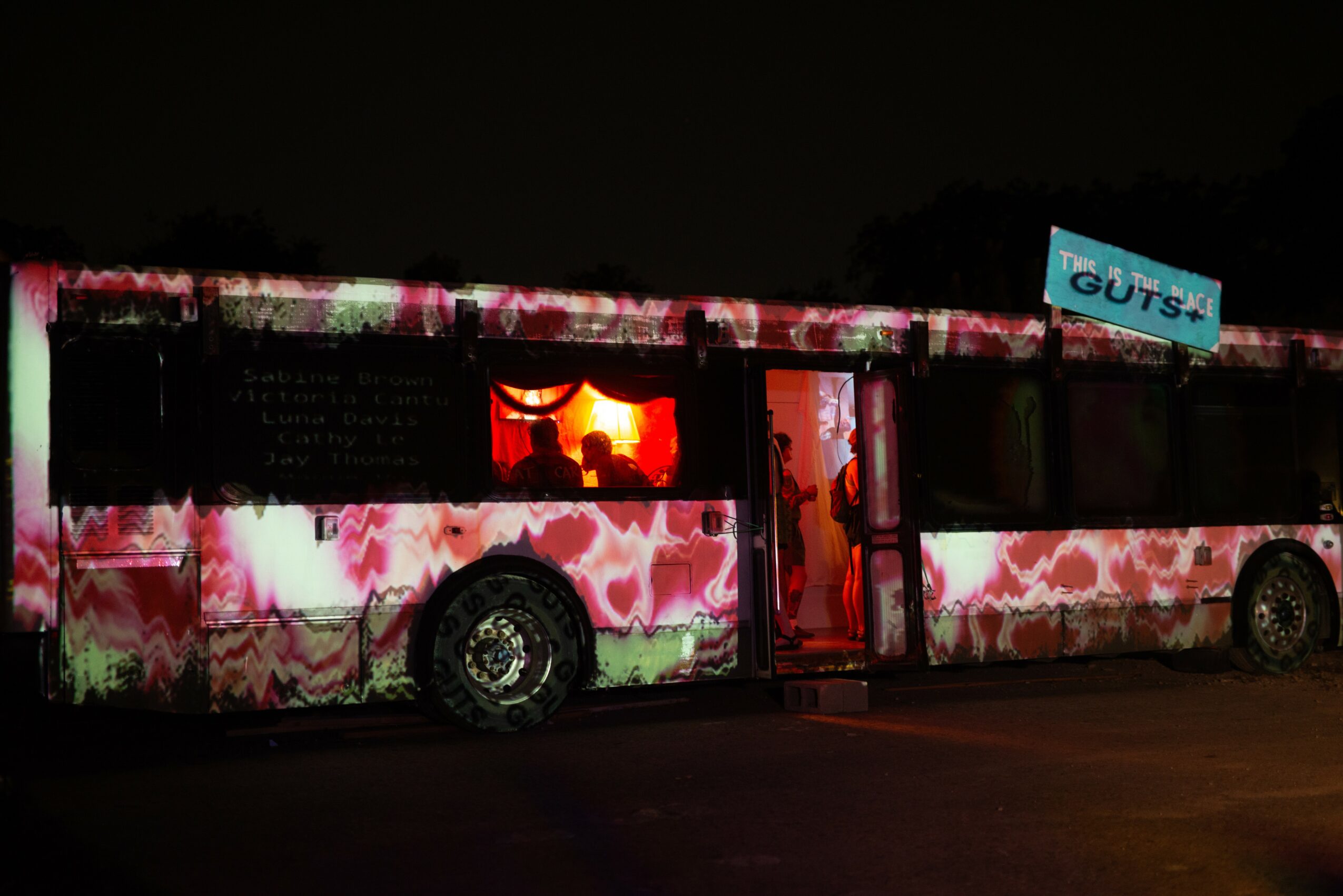 Bus with pink and white camouflage pattern, illuminated interior, and a blue sign on top, parked at night.