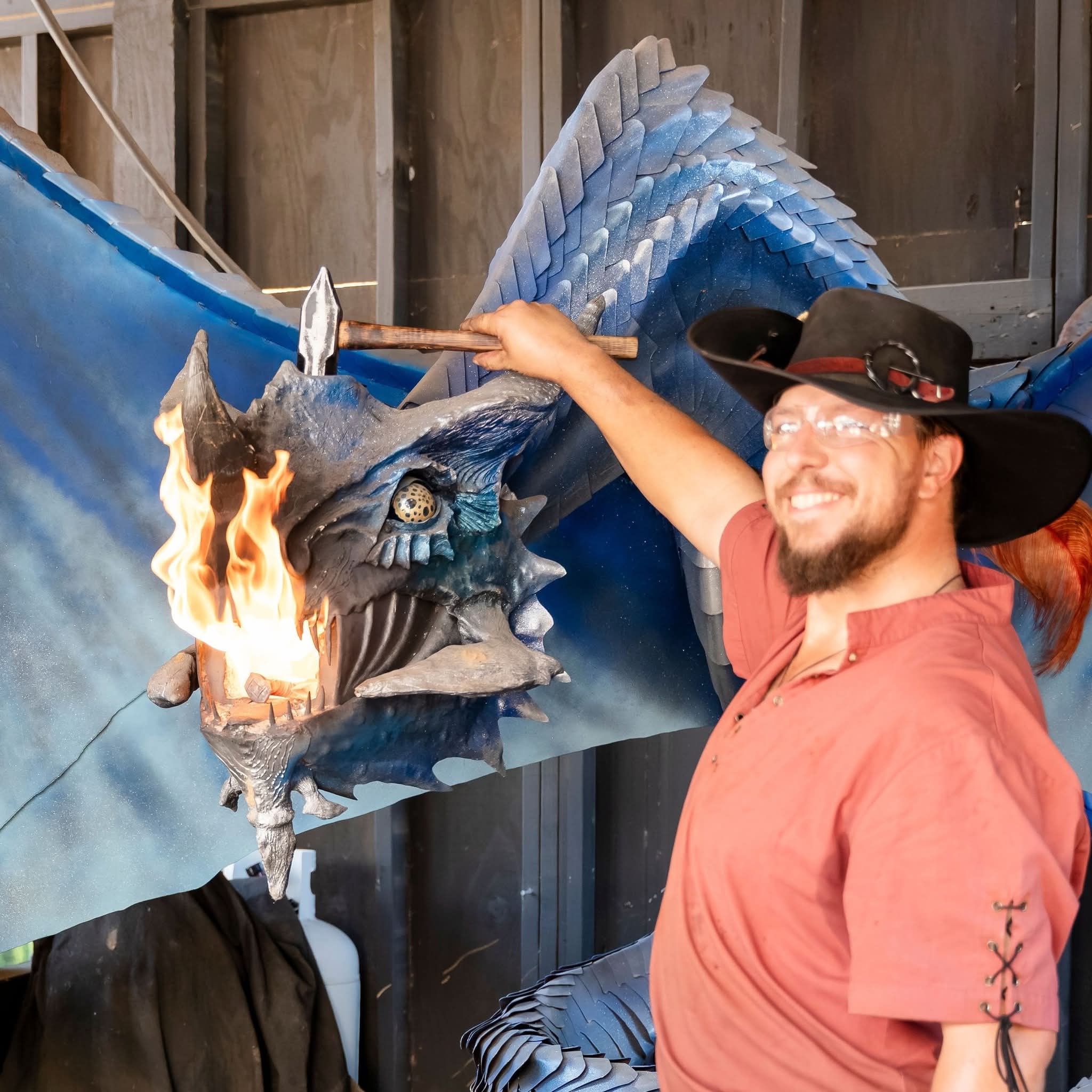 Man with glasses and a wide-brimmed hat smiling, touching a dragon sculpture with flames, in front of a wooden wall.