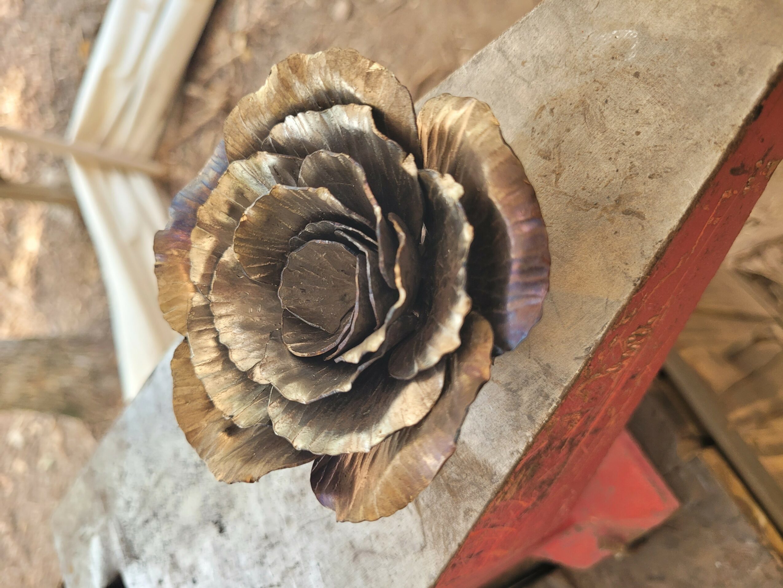 A metallic flower sculpture with layered petals, mounted on a concrete surface, with a blurred background.