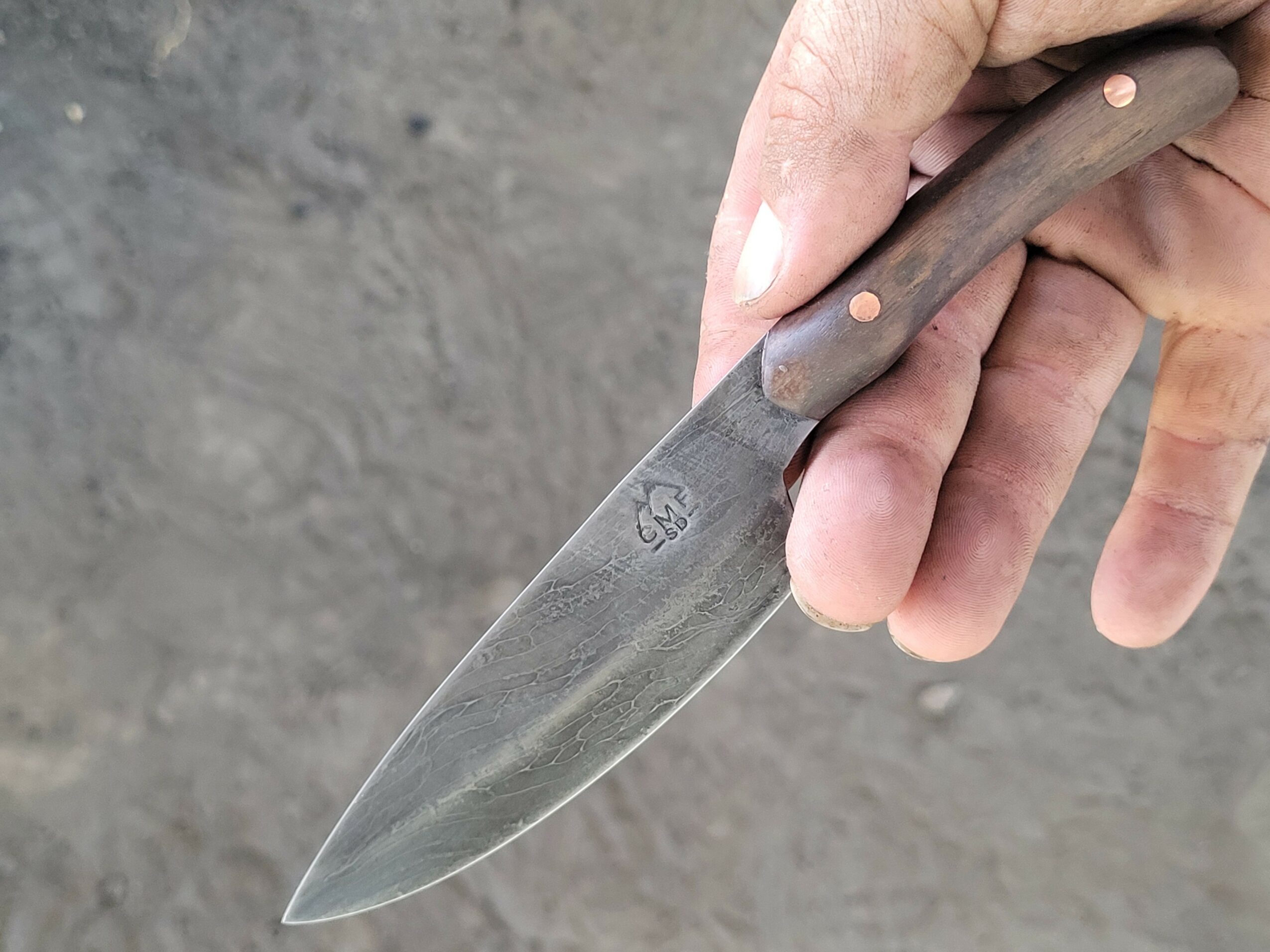 Hand holding a knife with a wooden handle, pointed downward, against a gray background.