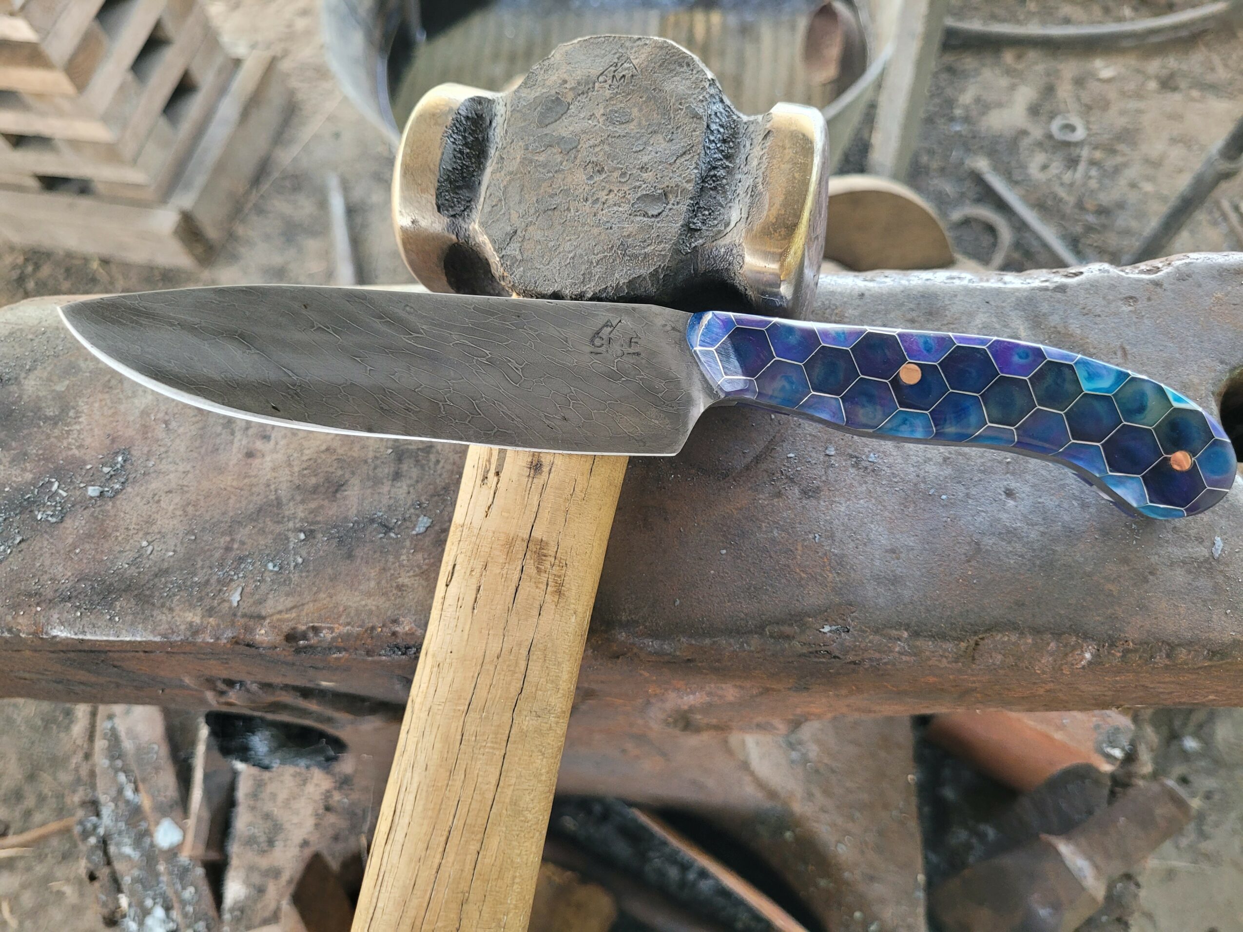 A knife with a blue hexagonal patterned handle resting on a workbench, with a hammer and wooden block nearby.