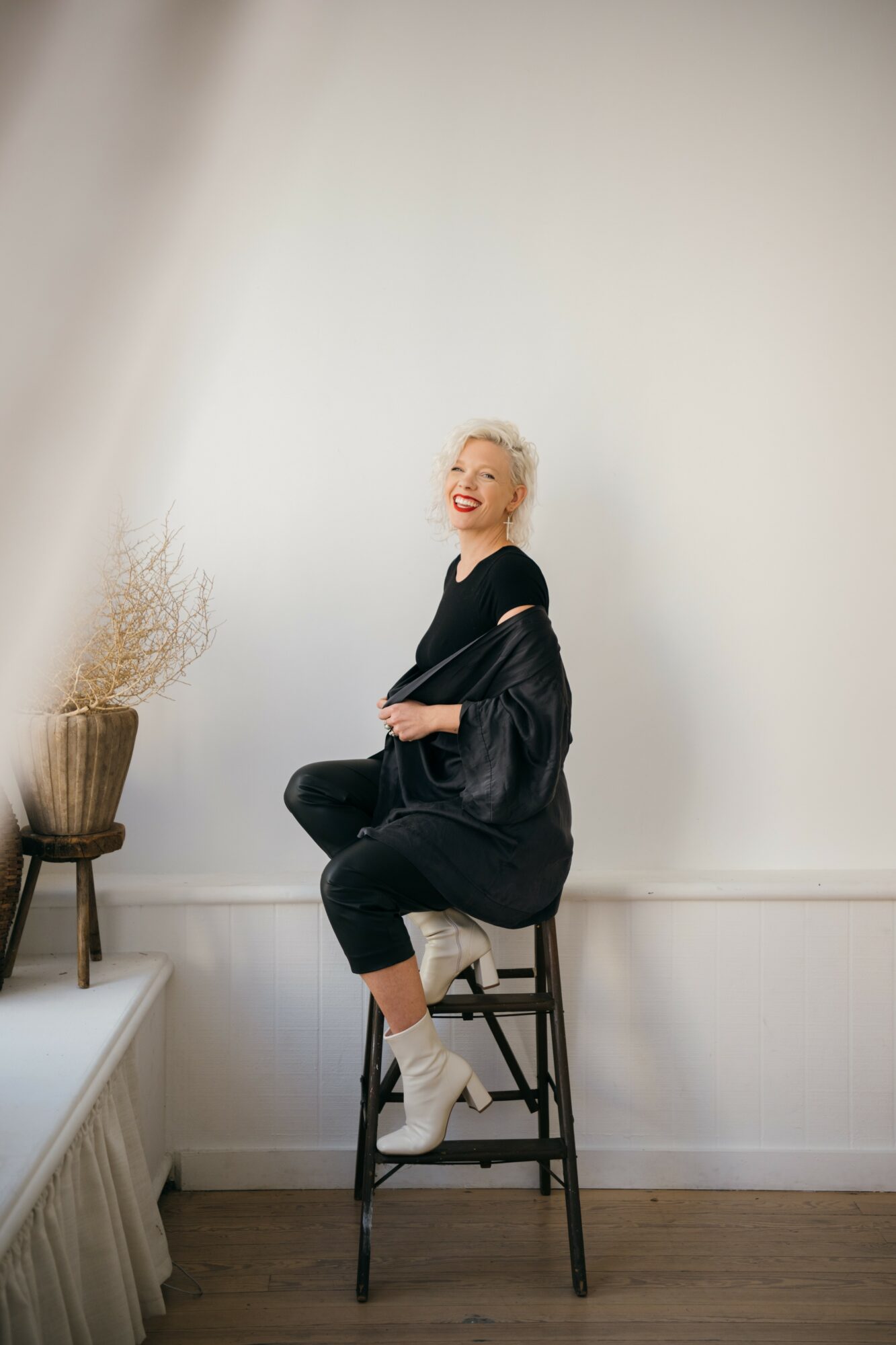 Woman with short blonde hair smiling, sitting on a stool in a minimalistic room, wearing black clothing and white socks.