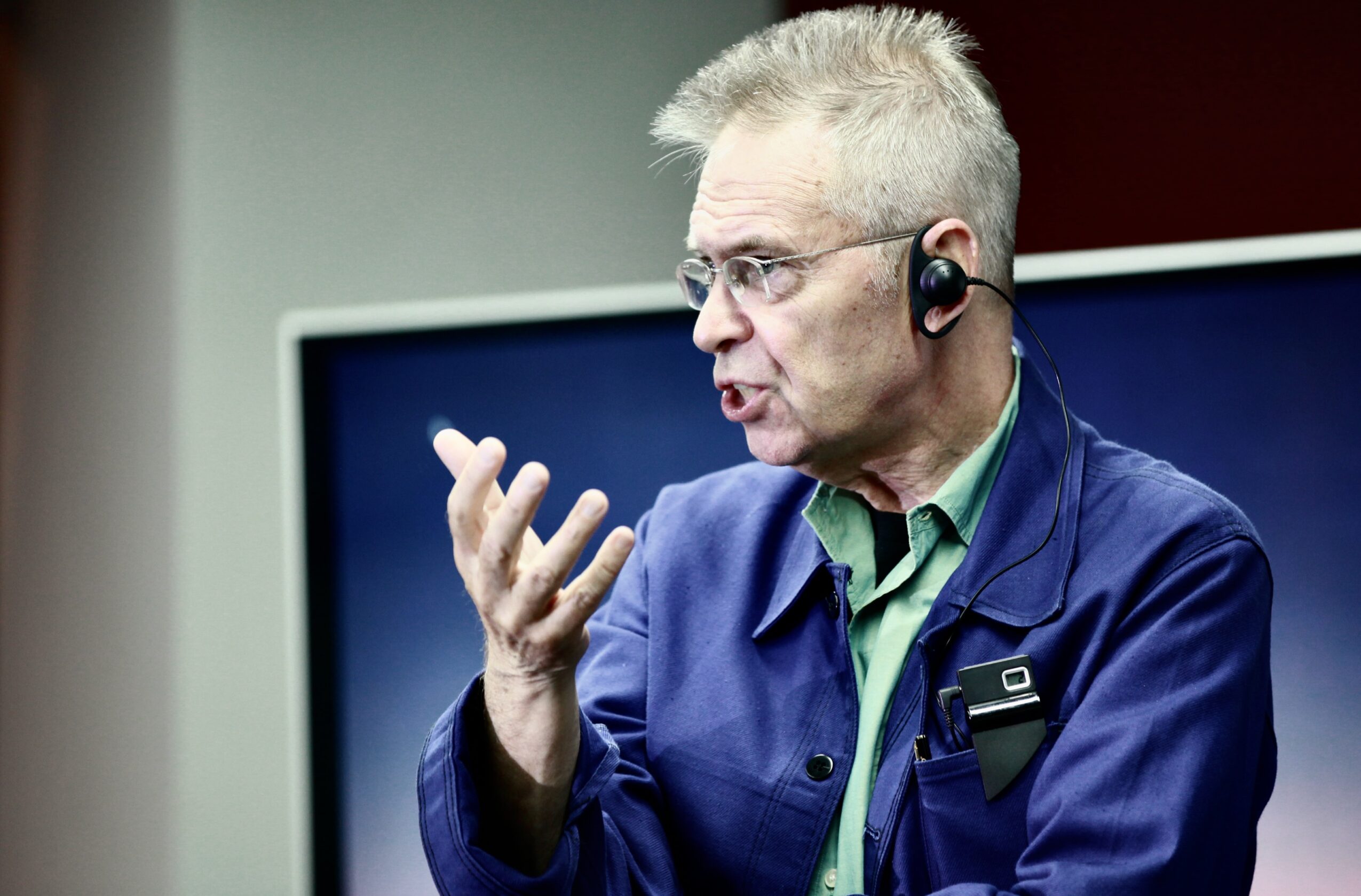 Elderly man with glasses and short gray hair speaking, wearing a blue jacket and headset, in front of a screen.