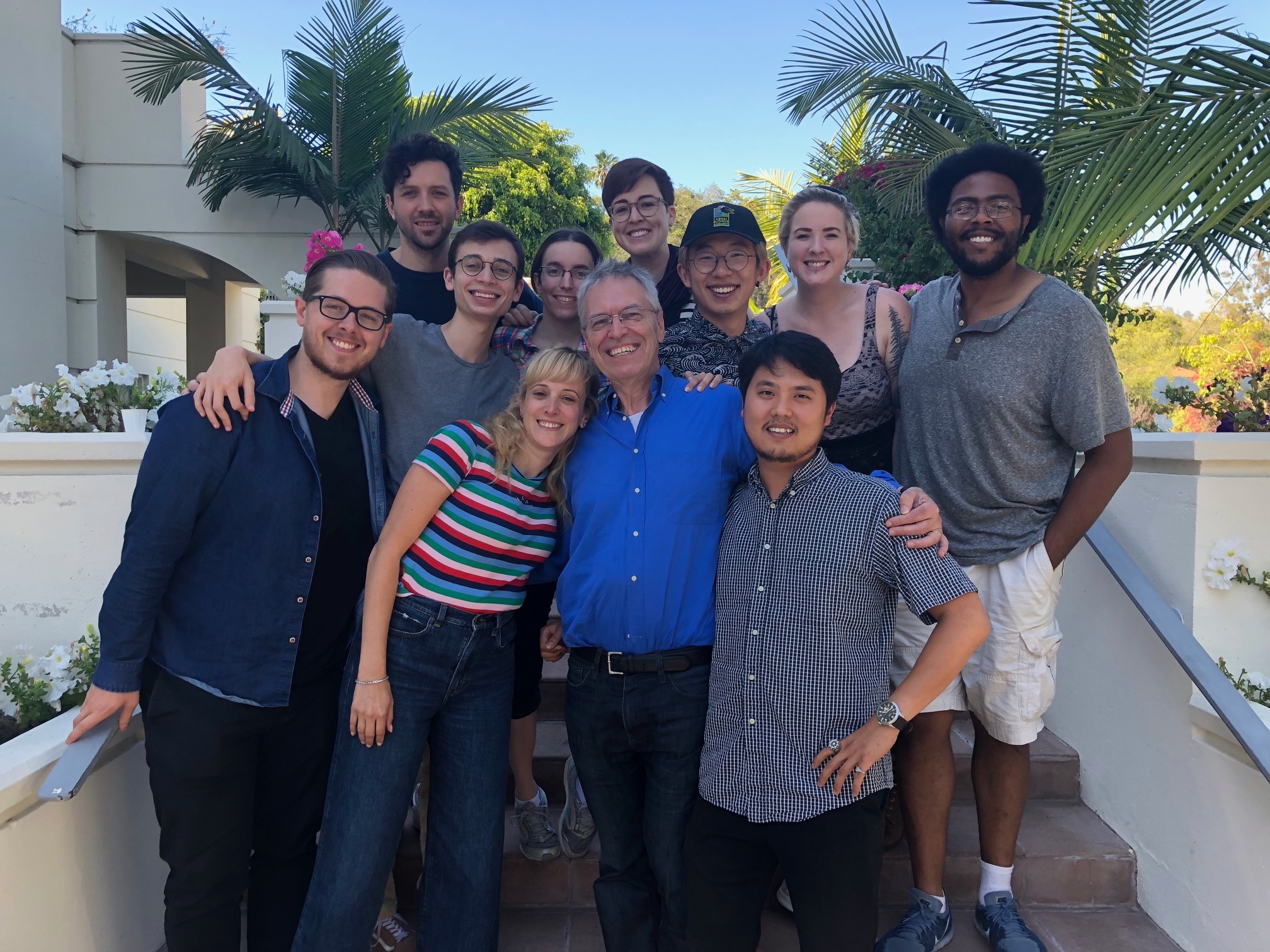 Group of ten people smiling outdoors with palm trees and a building in the background.