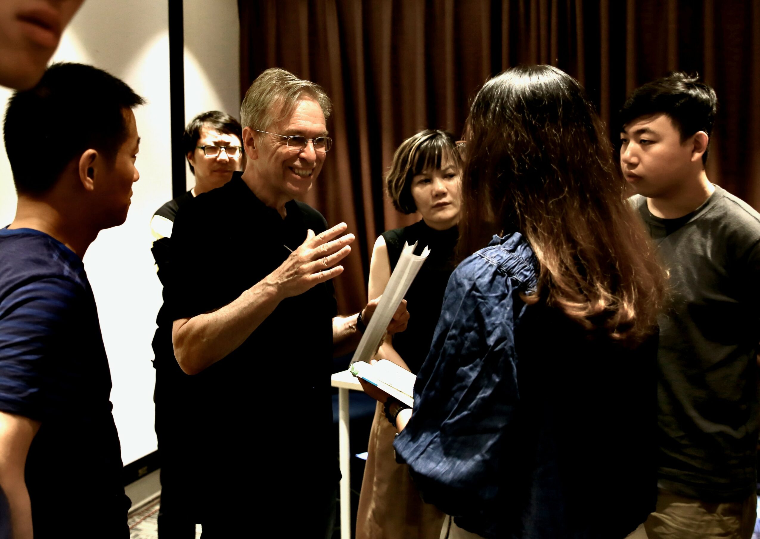 Group of people, including a smiling man with glasses, engaged in conversation indoors with brown curtains in background.