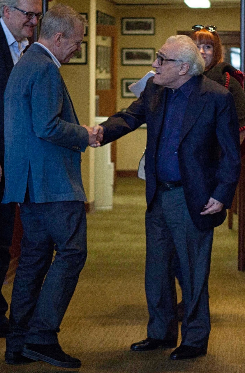 Two men shake hands in a room with framed pictures on the wall, one wearing a blue suit and glasses, the other in a dark suit.