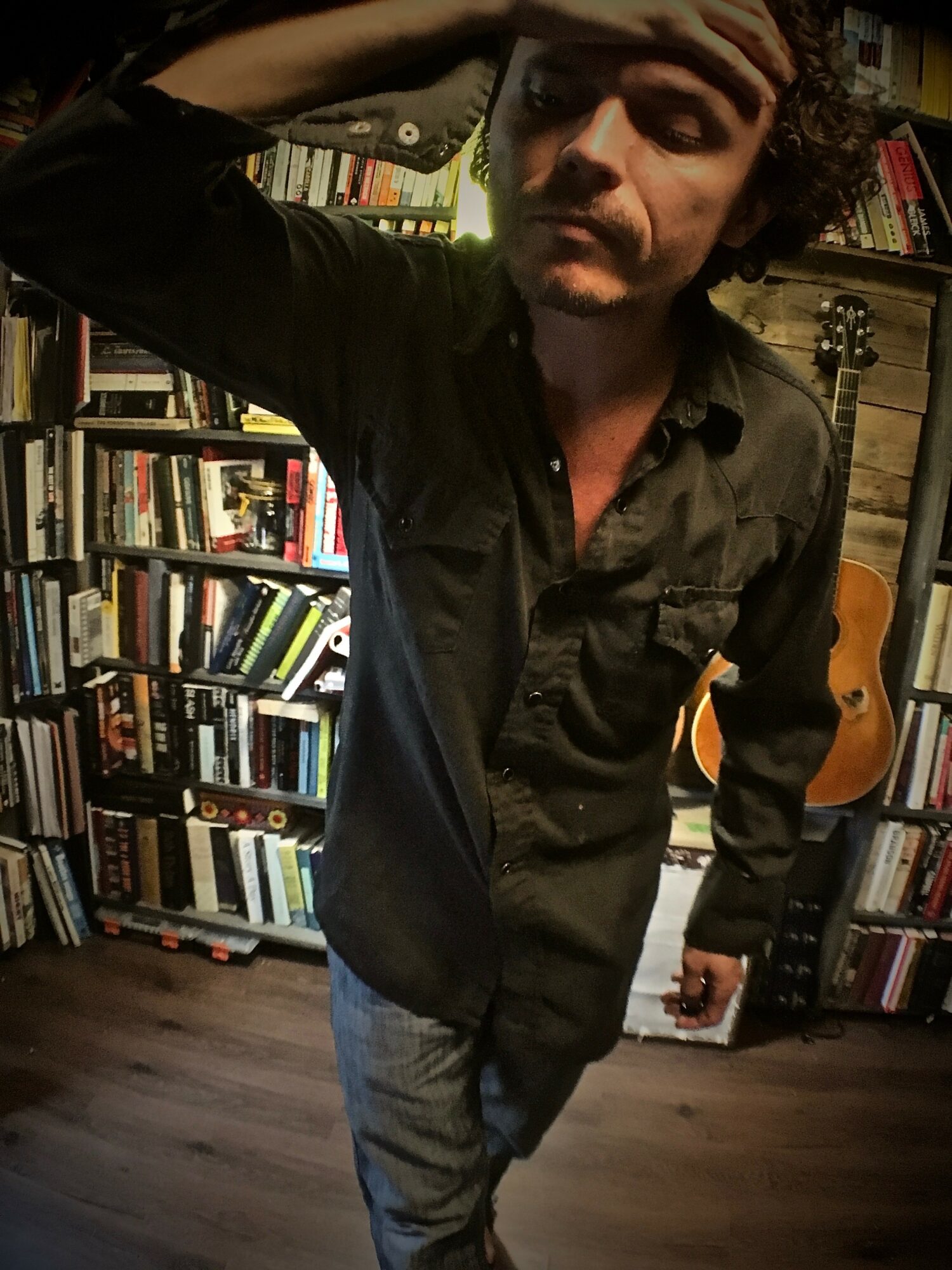 Young man with curly hair and a dark shirt stands in a room with bookshelves and a guitar, touching his forehead.