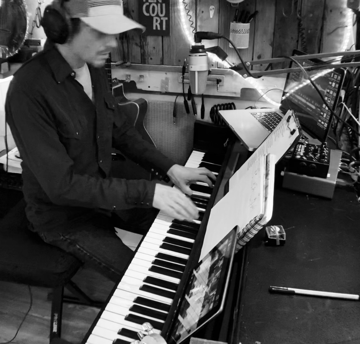 Person playing piano with sheet music, wearing a cap and headphones, in a studio setting.