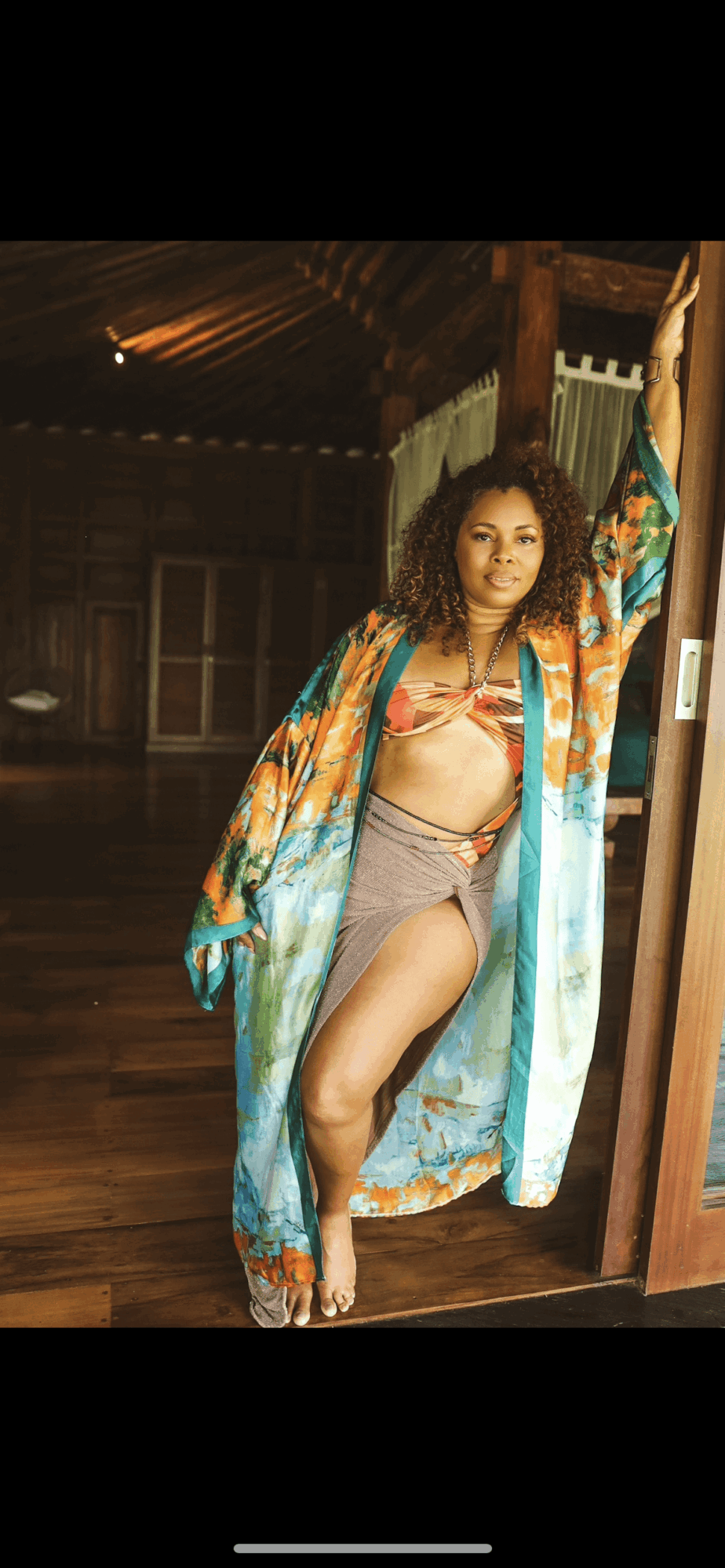 Woman with curly hair in a colorful kimono and swimsuit standing in doorway, wooden interior background.