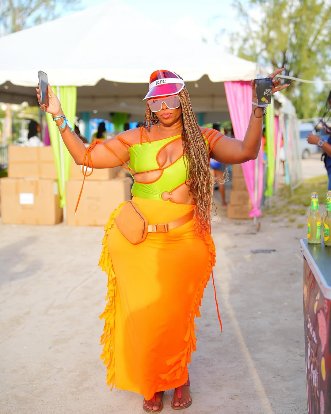 Woman in colorful outfit holding two sticks at outdoor event, with tents and trees in background.
